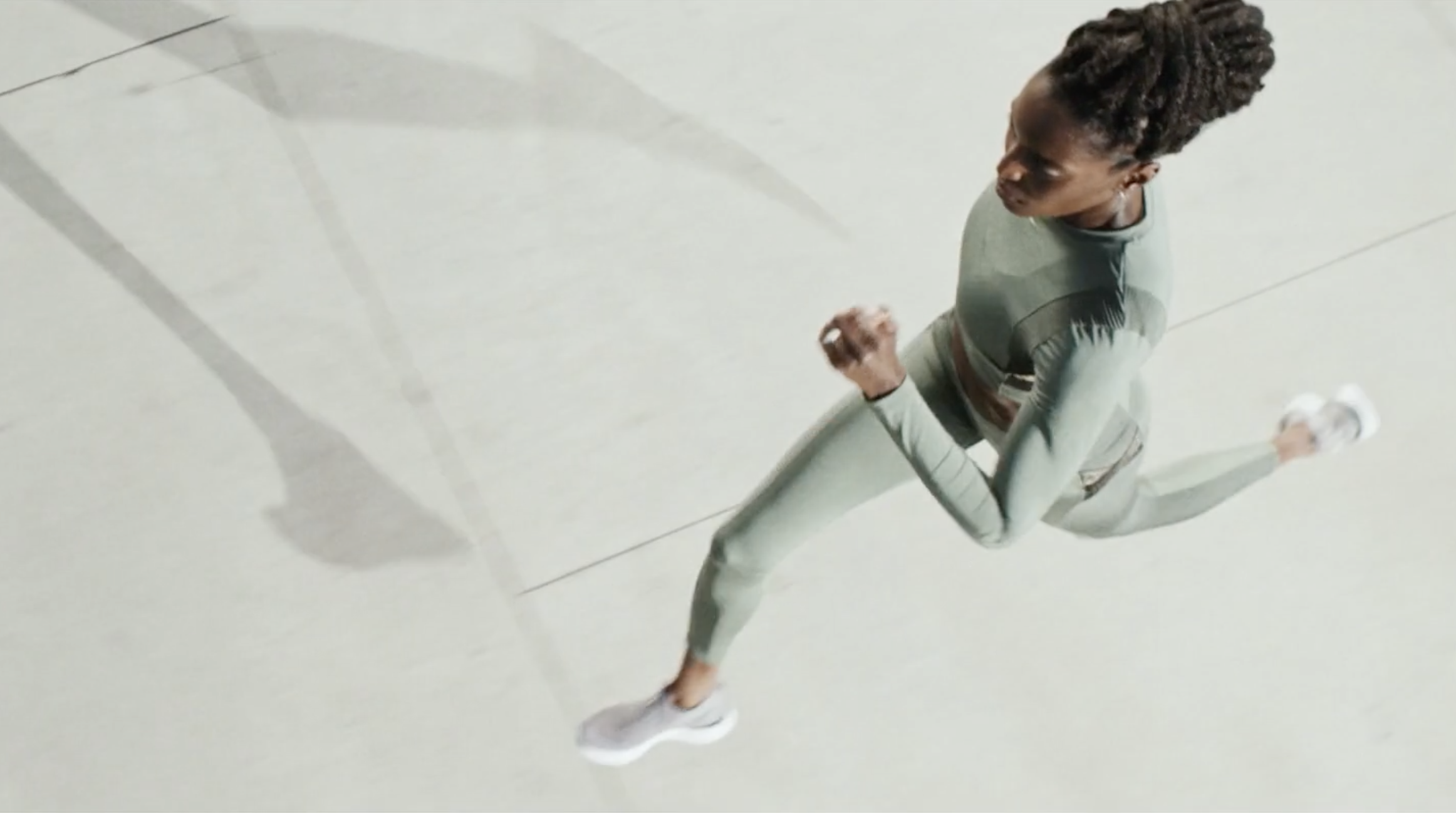 An aerial view of a woman running on the floor, wearing a gray sports outfit and white sneakers, casting a shadow.