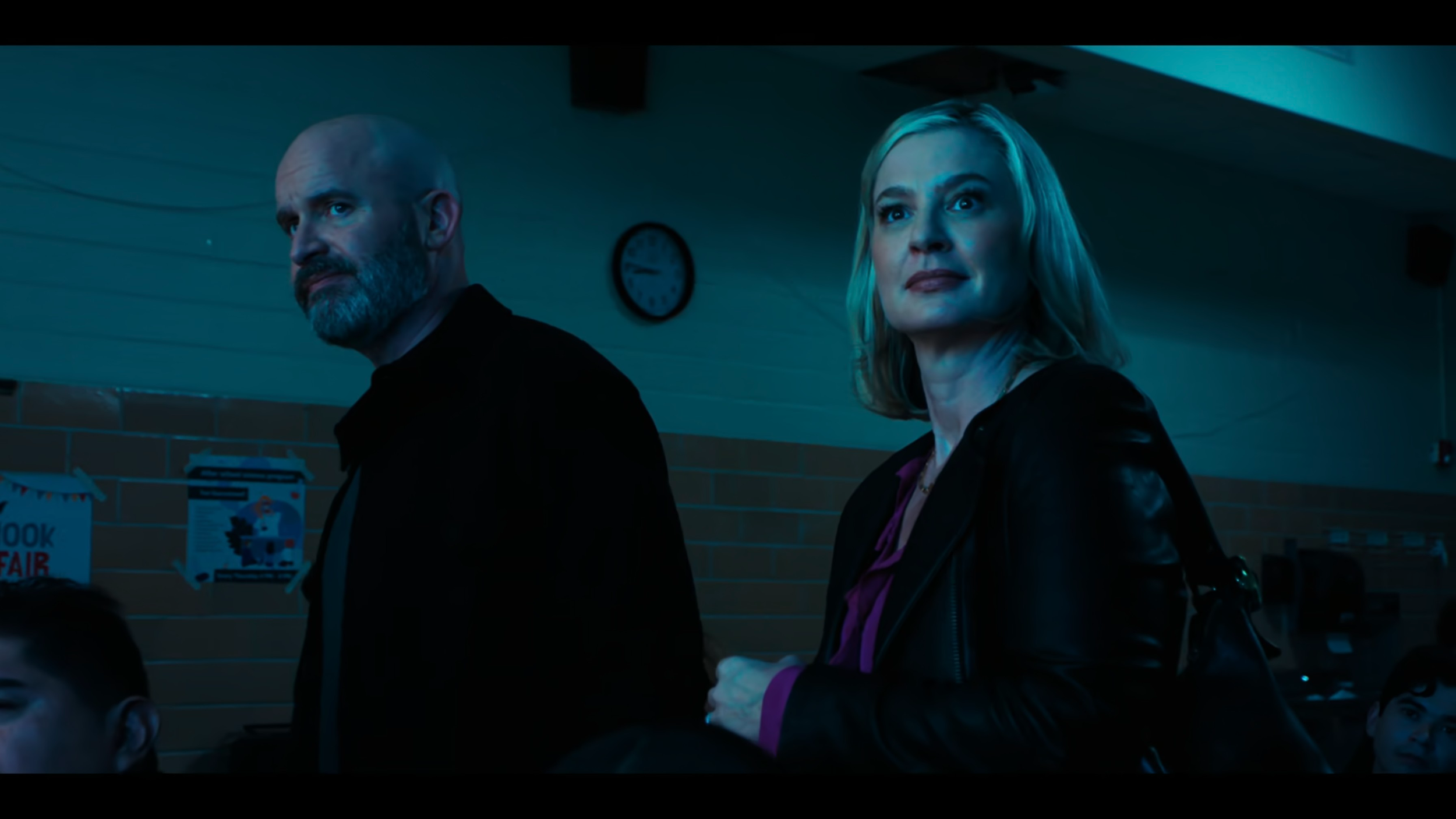 A man with a bald head, gray beard, and black clothing standing next to a woman with blonde hair, wearing a black leather jacket and purple blouse, in a dimly lit room with a clock showing 8:20 on the wall.