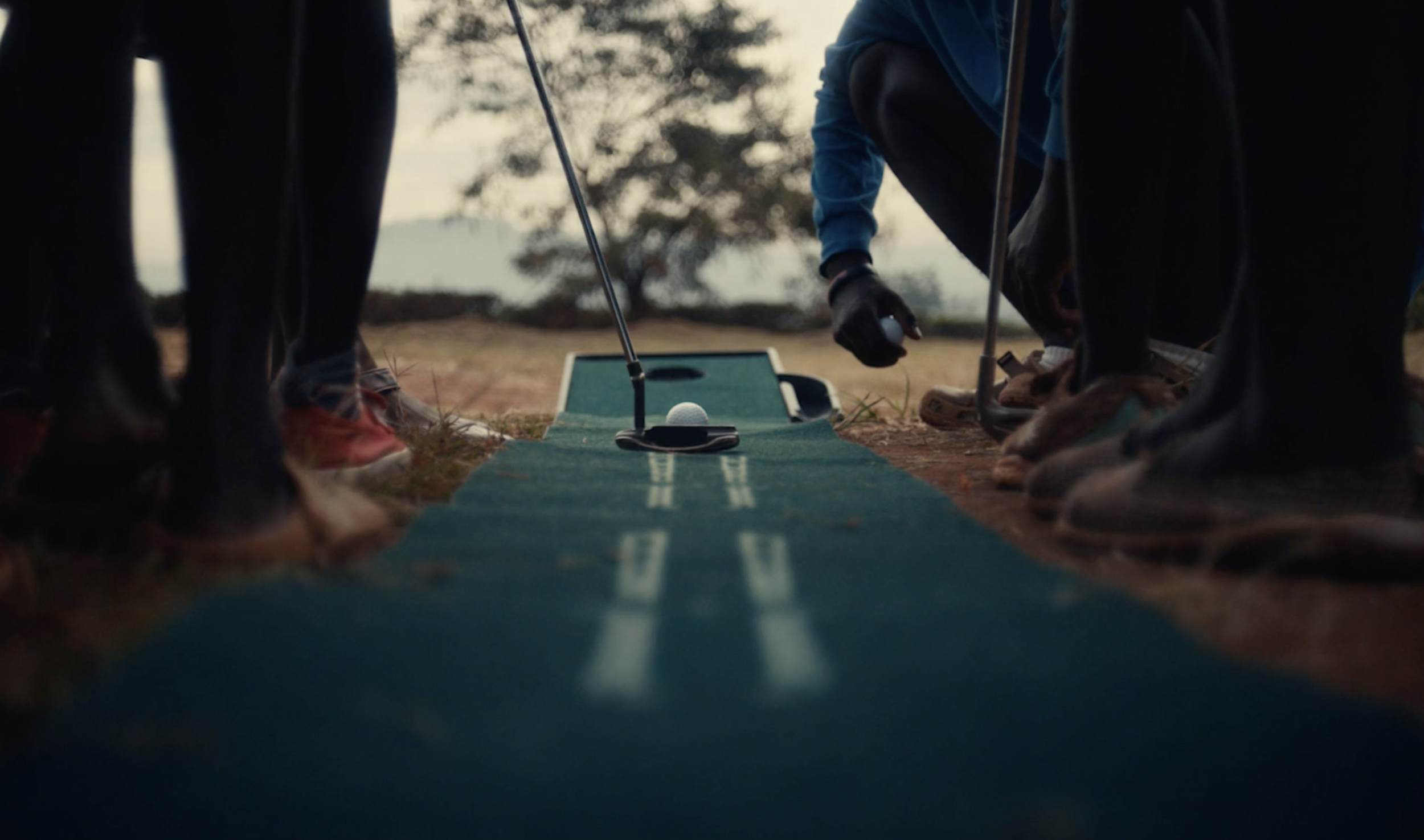 Close-up view of a golf course with a putter, golf ball, and a putting green, surrounded by several people preparing to practice putting outdoors during sunset.
