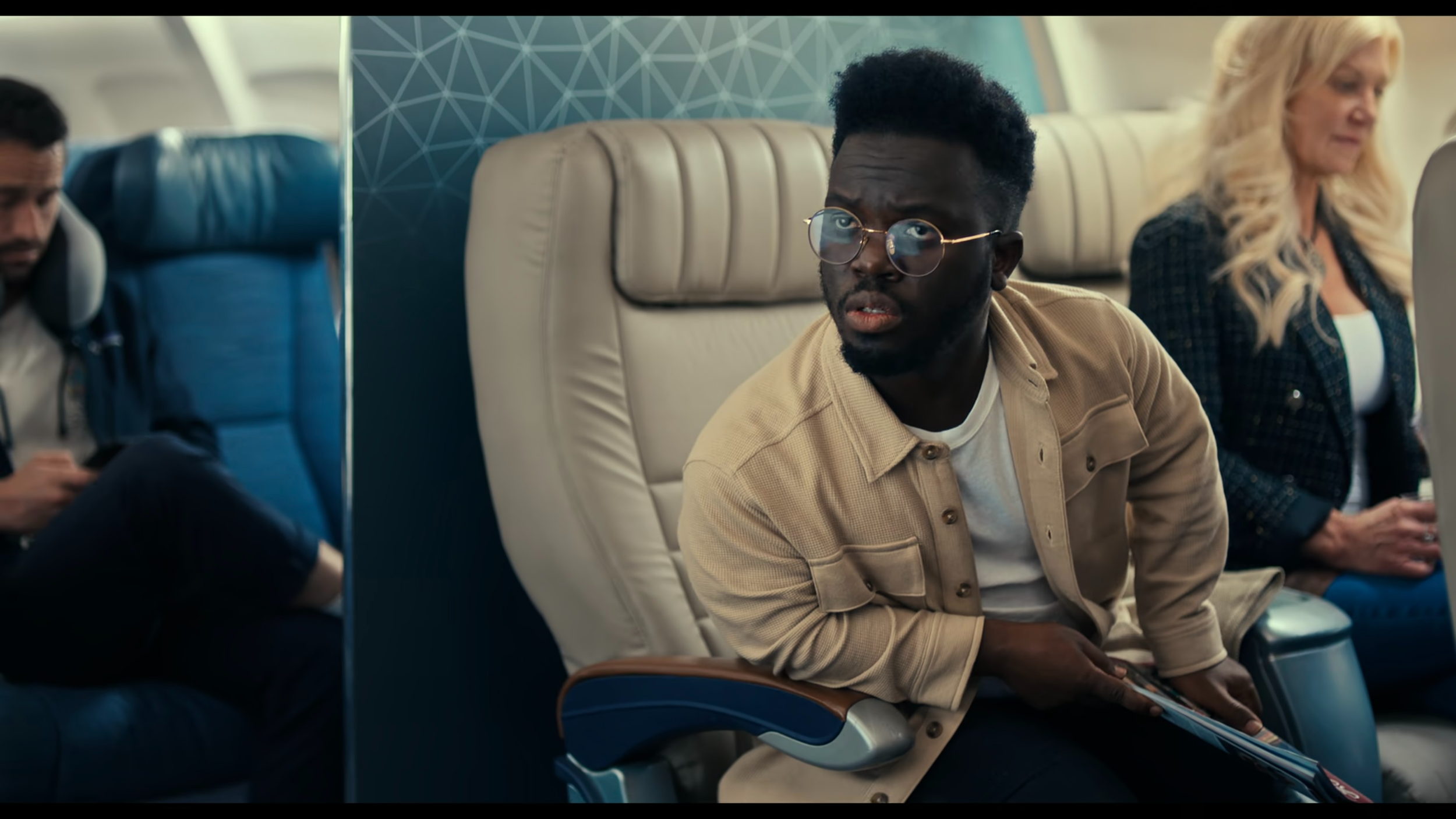 A man with glasses and a beige jacket sitting in an airplane seat, looking surprised or confused. There are two other passengers visible in the background, one man to the left using a phone and a woman to the right reading.