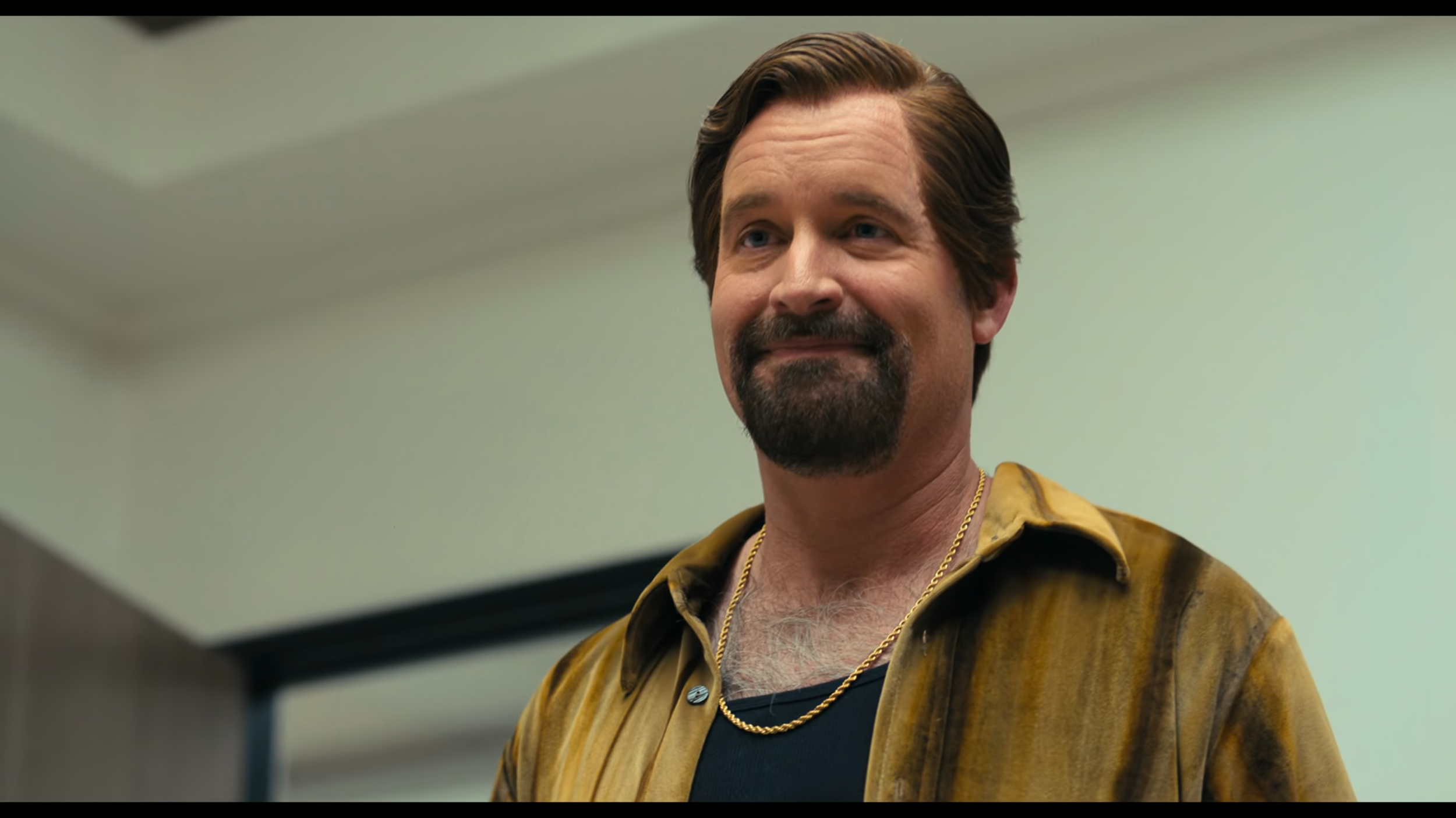 A man with a beard and mustache, wearing a gold chain and a yellow-brown shirt, smiling in a room with a pale green wall.