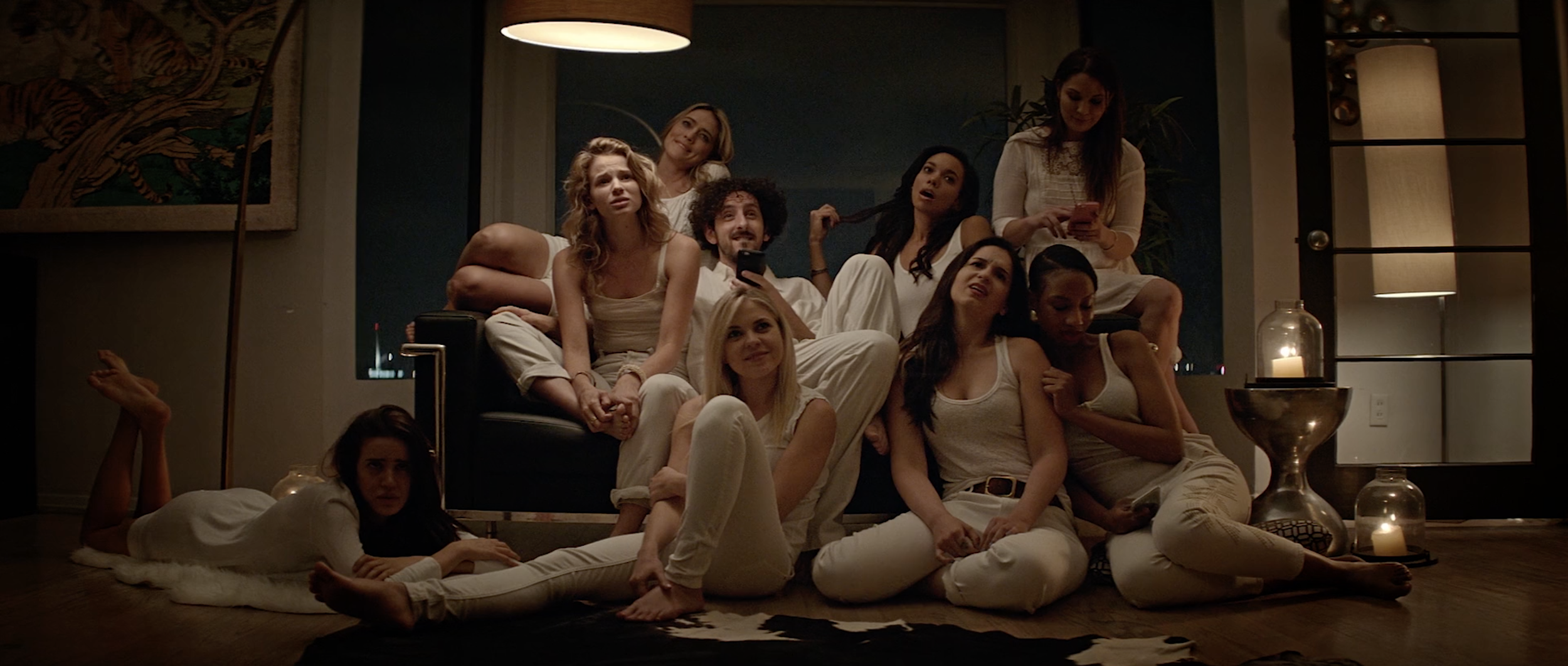 A group of nine women are gathered in a living room, sitting on a sofa and the floor, in a relaxed, intimate setting. They are dressed in white or light-colored clothing, with some holding phones. The room has dim lighting with candles and a lantern on the right, and a modern decor with a wall art piece on the left.