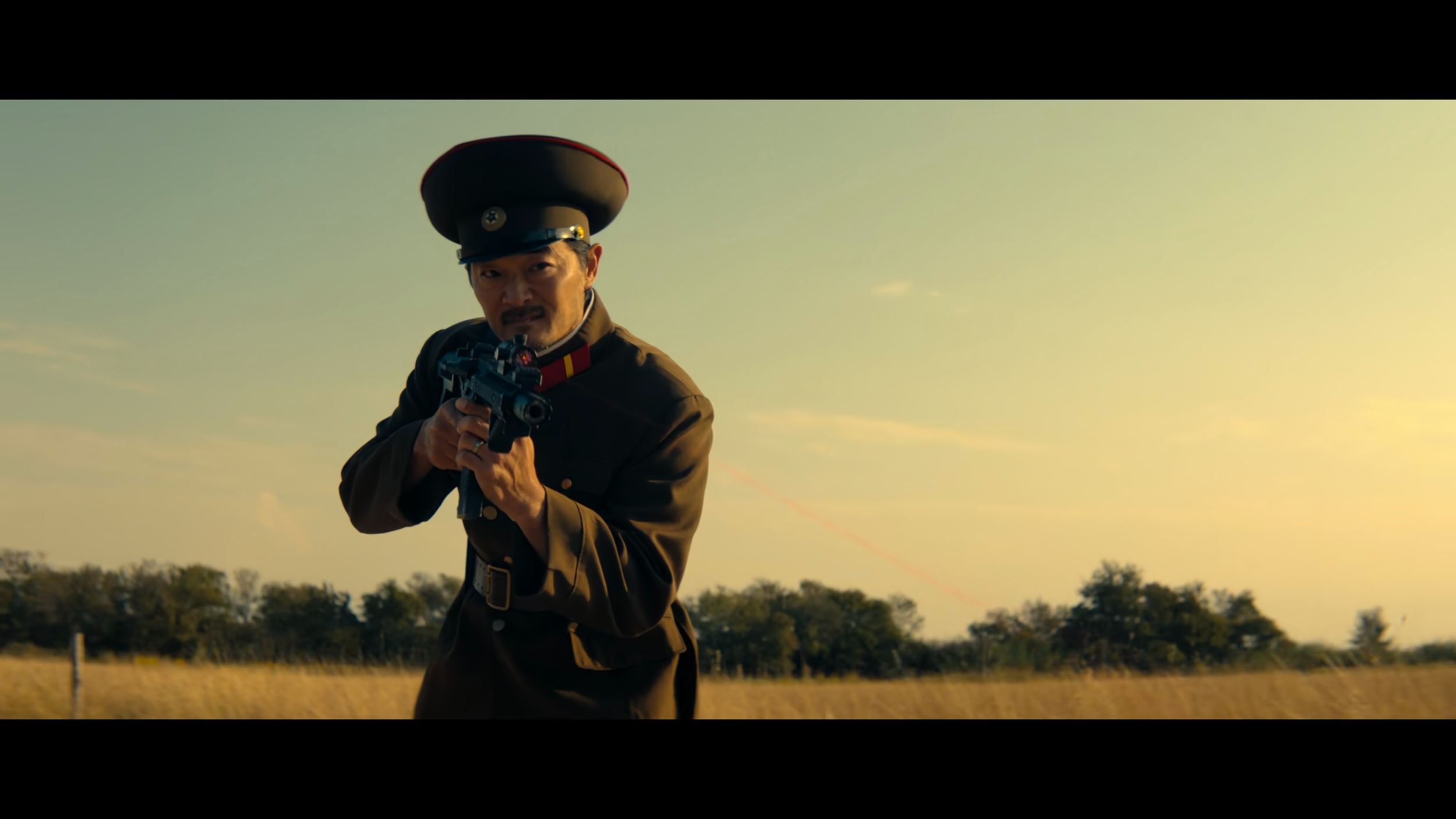 A man dressed in a vintage military uniform with a peaked cap, aiming a pistol outdoors at sunset, against a background of trees and open field.