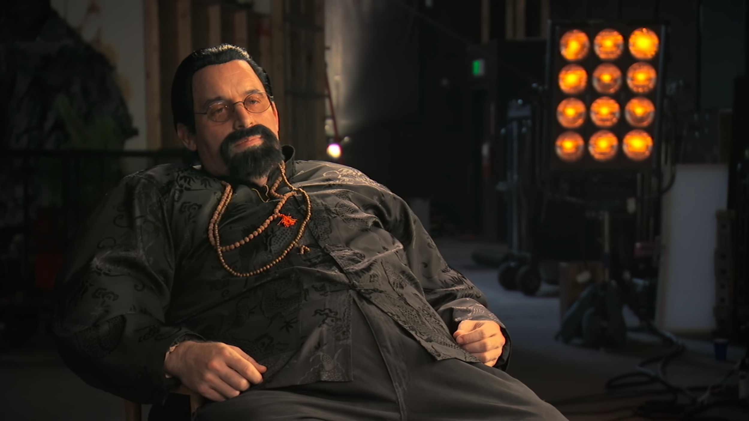 A man with glasses and a beard, dressed in a dark patterned shirt, is sitting and looking relaxed in a dimly lit indoor setting with stage lights and equipment in the background.