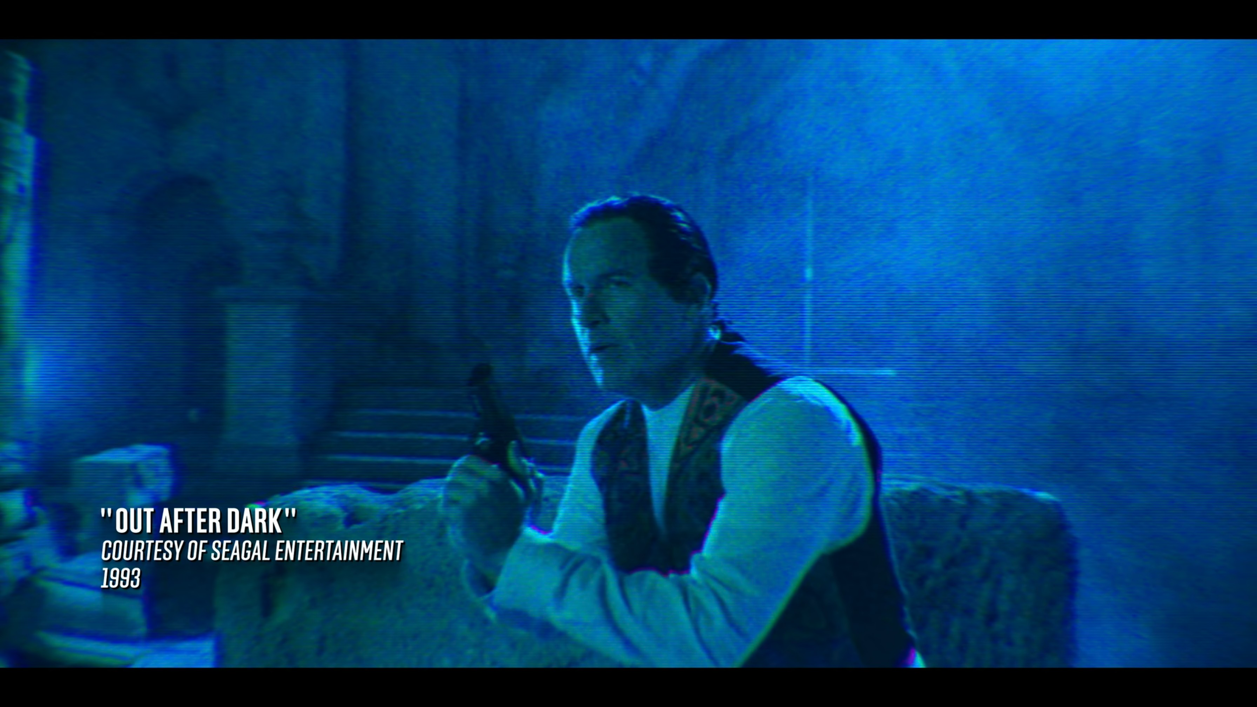 A man sitting at a table in a dimly lit, blue-toned scene, holding a gun near his face, with text overlay indicating the movie title 'Out After Dark,' in 1993, courtesy of Seagal Entertainment.