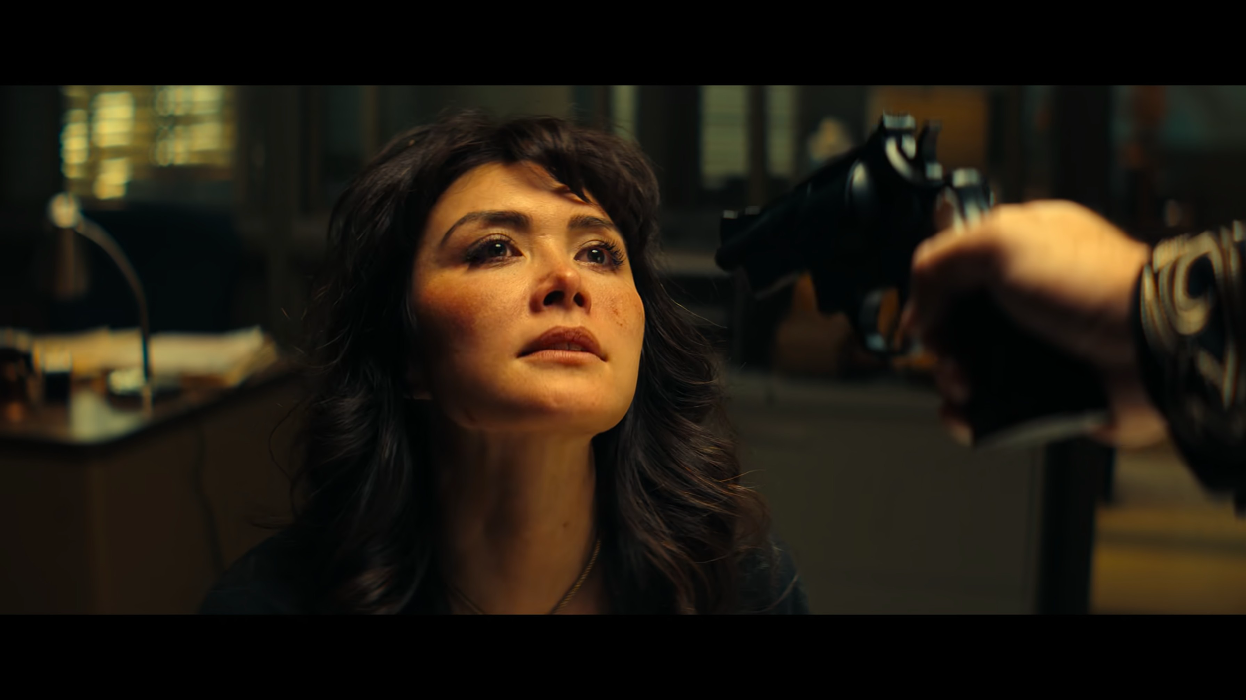 A woman with dark curly hair looking slightly upward as a gun is pointed at her head.