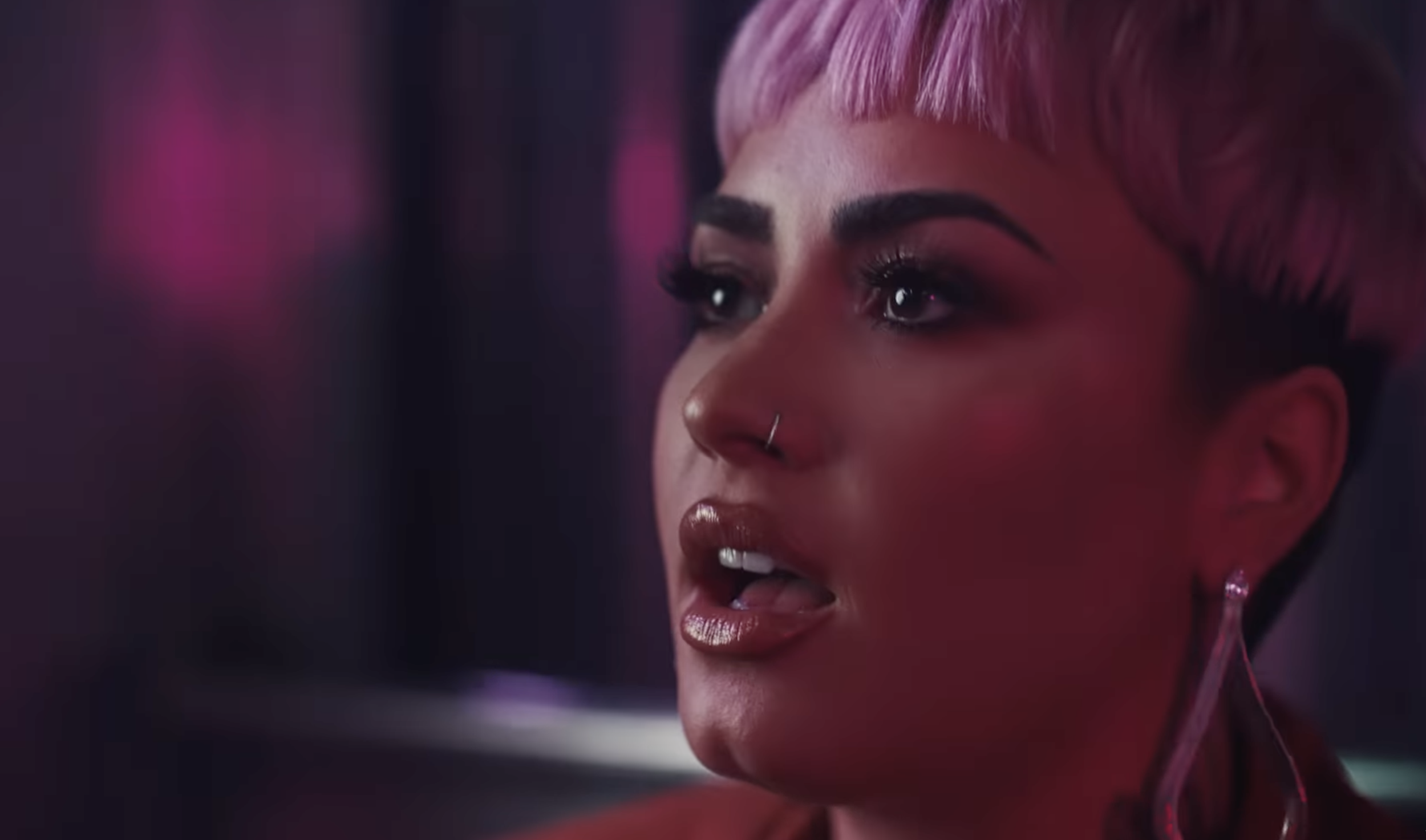 A woman with blonde hair and a nose ring, wearing makeup and earrings, making a playful face with her tongue slightly out, in a dimly lit environment with pink and purple lighting.