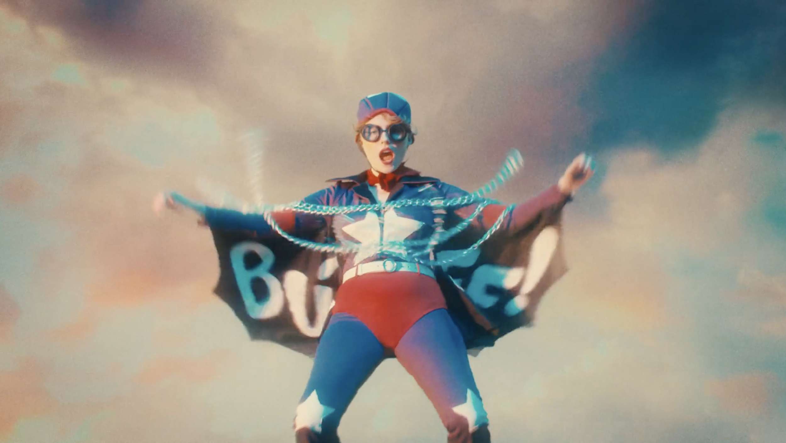A person dressed in superhero costume with a cape and mask, standing against a cloudy sky, with arms outstretched.