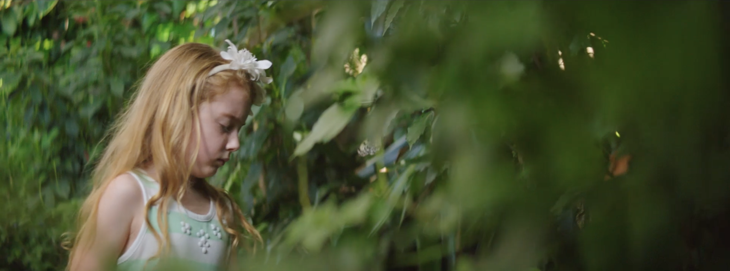 A young girl with long red hair wearing a white dress with light green accents and a floral headband, standing amidst green foliage.