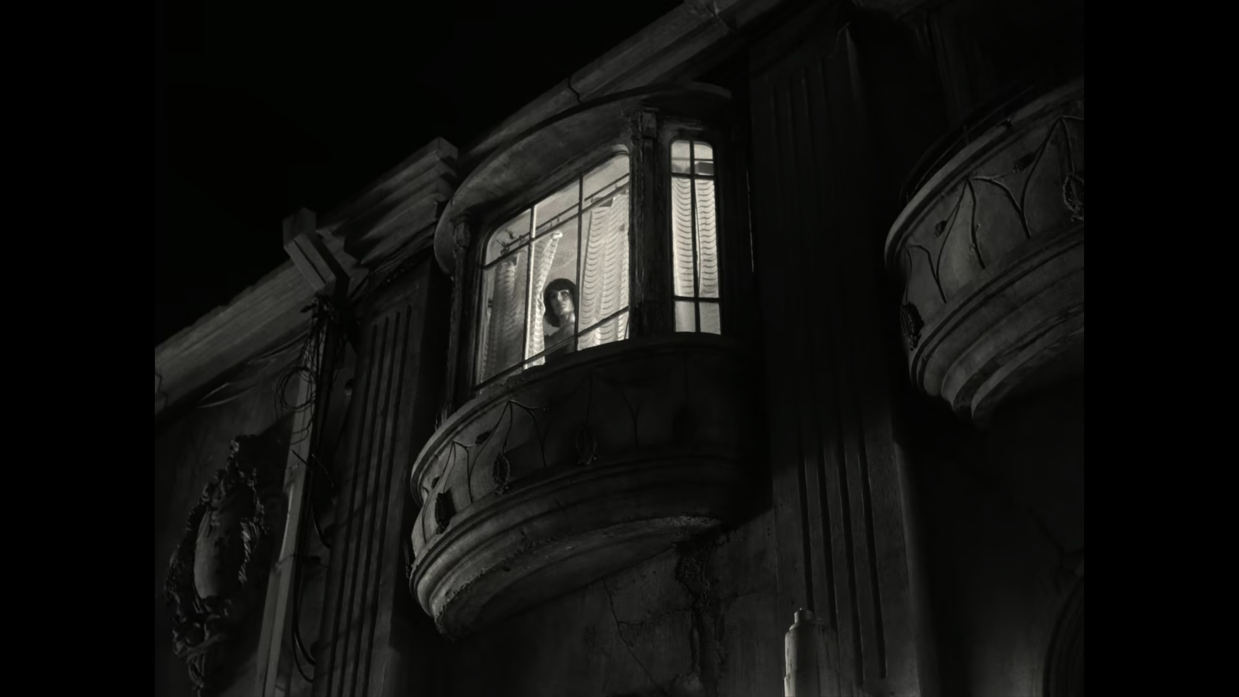 A woman looking out from a tall, narrow window with curtains in an old, dark building or house at night.