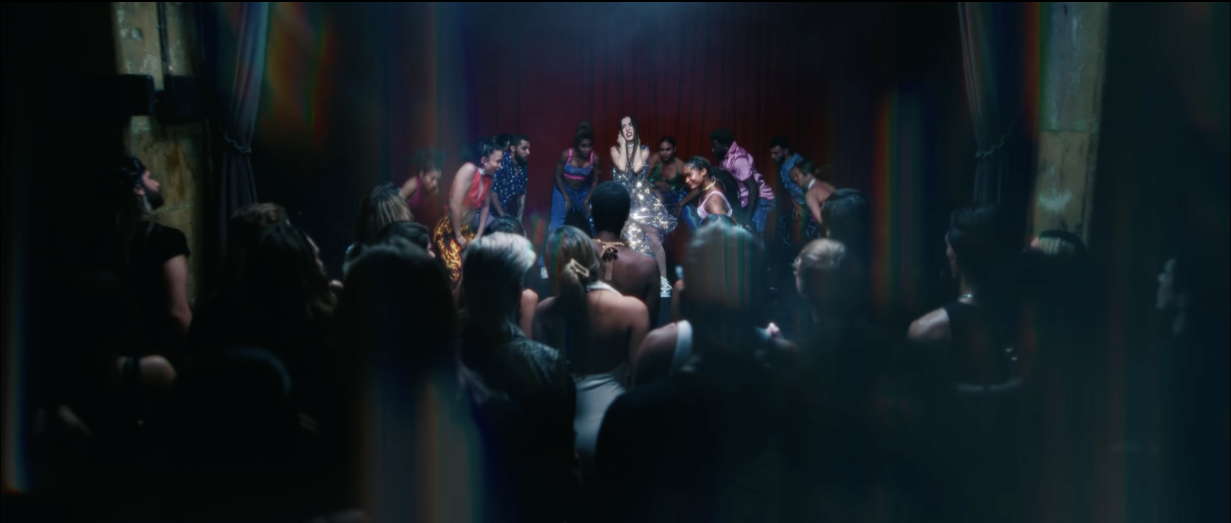 A woman in a shiny dress performs on stage in front of an audience while dancers surround her.