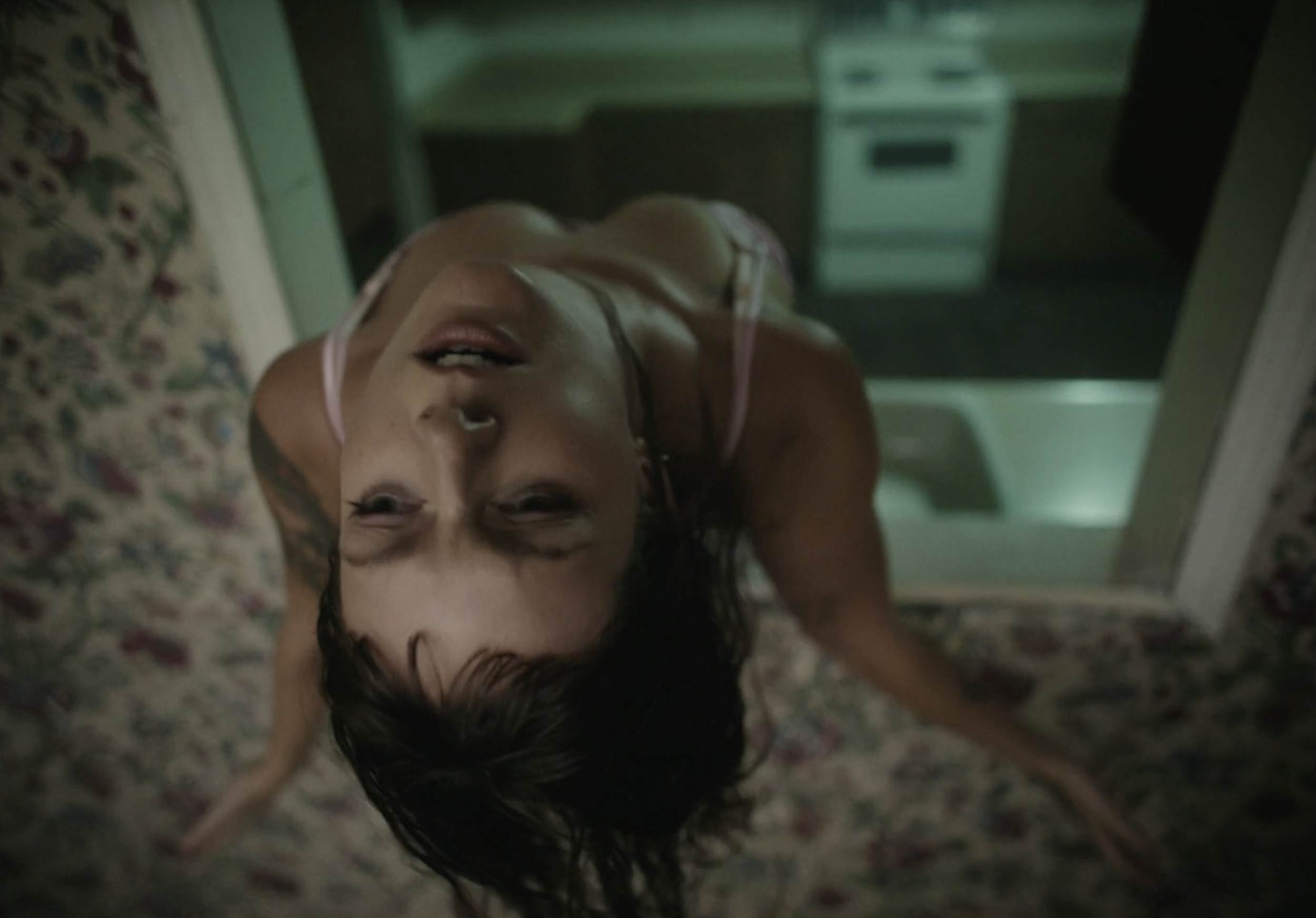 A woman with wet hair and tattoos on her arms is lying on her back on a floral carpeted floor. She is wearing a pink spaghetti strap tank top. The photo is taken from above, with the woman looking up at the camera.