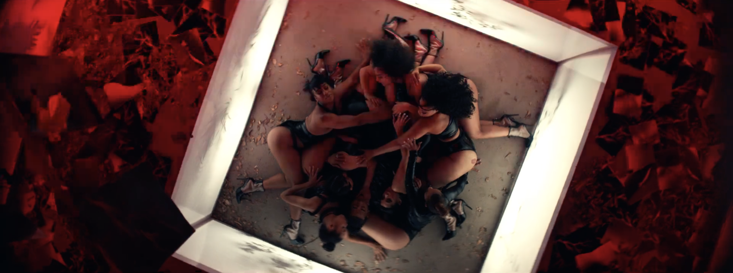 Six women dressed in black outfits and high heels are wrestling on the floor inside a white frame, with red abstract background surrounding it.
