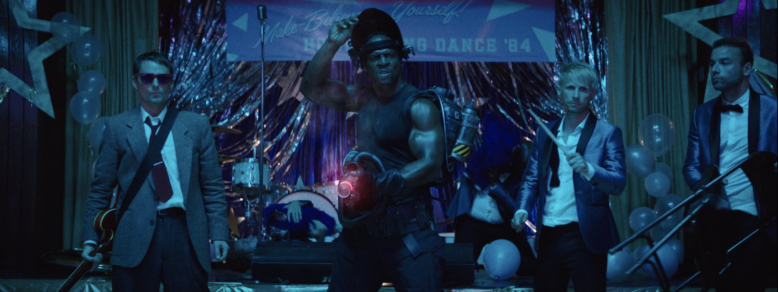 A group of four men in suits holding musical instruments and a chain saw on a stage decorated with balloons and a shiny backdrop, with a banner reading 'Make Believe Yourself'.