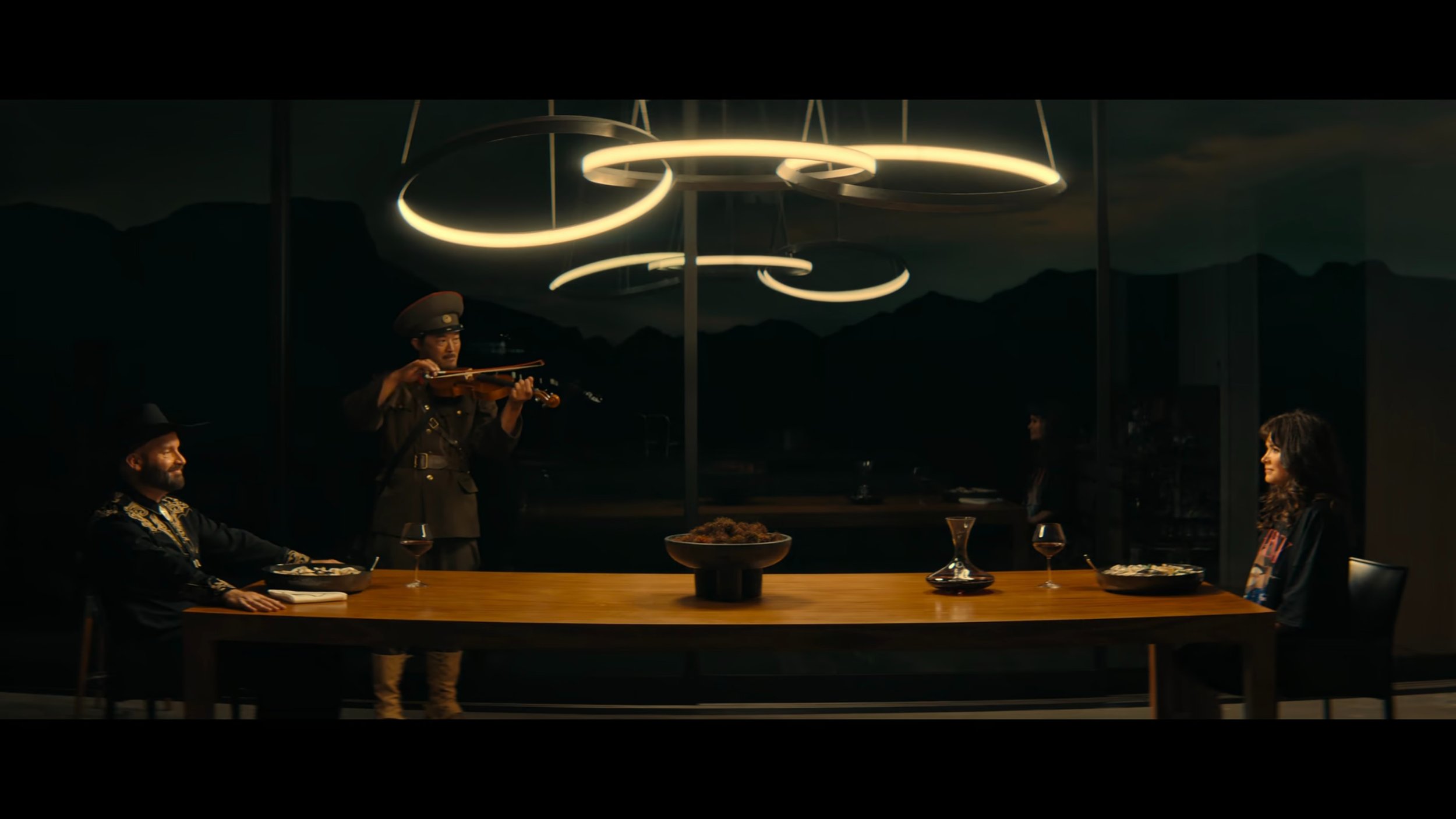 People dining at a table with a violinist playing in front of a glass window, dark mountains in the background, modern circular ceiling lights above.