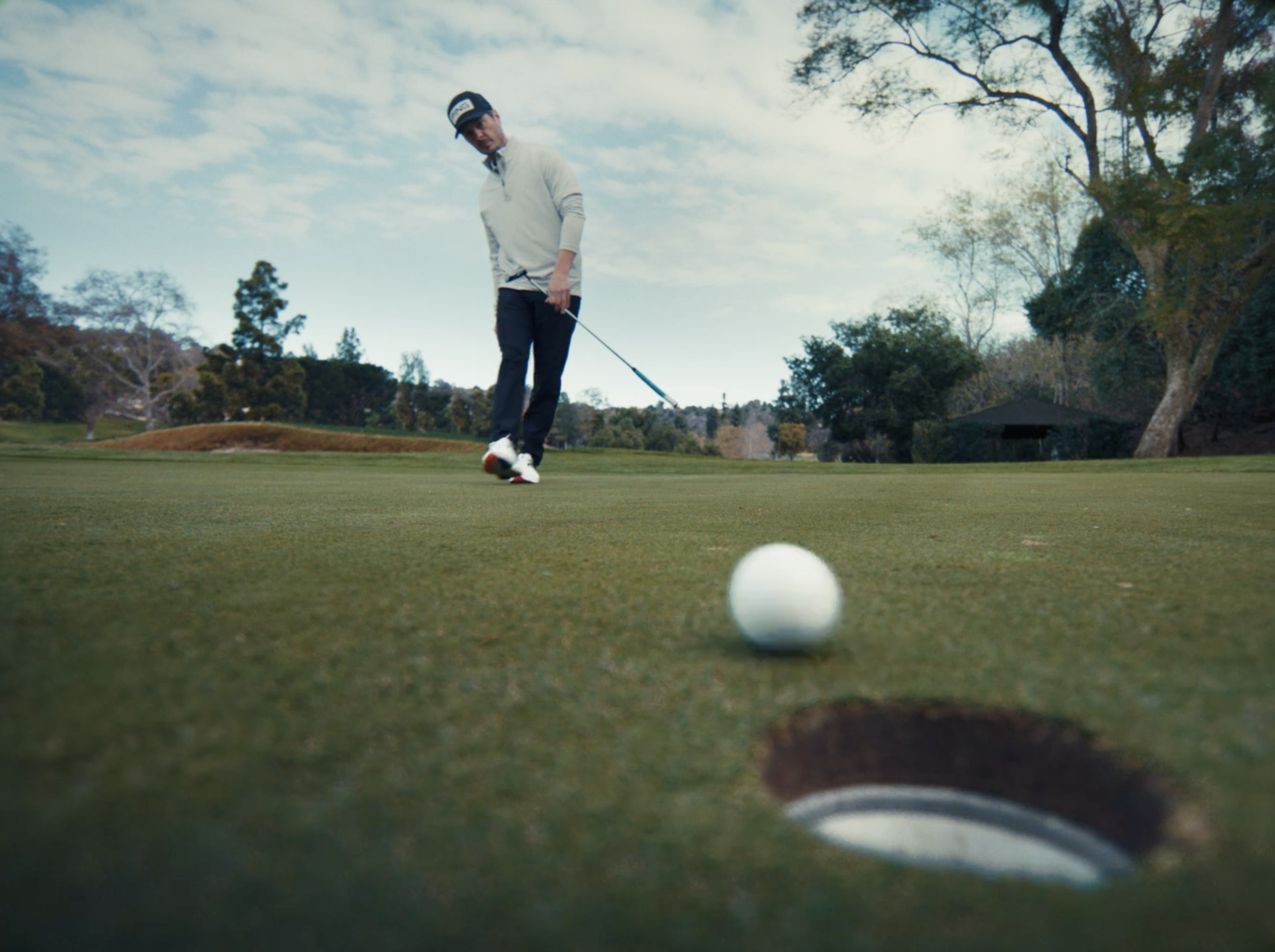 A man preparing to hit a golf ball on a golf course, with a hole and flag in the foreground, trees, and a cloudy sky in the background.