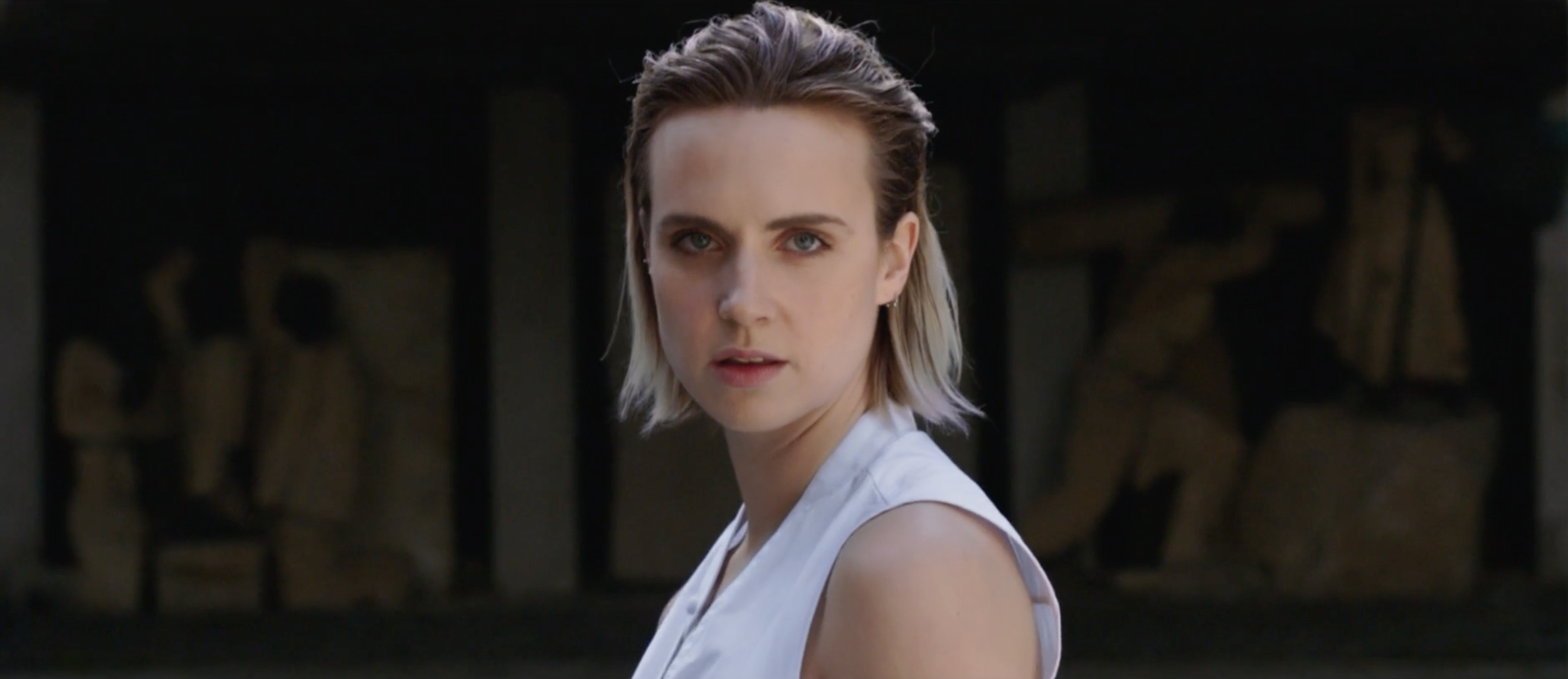 A woman with short blonde hair and blue eyes looking over her shoulder in front of a background of wooden logs.