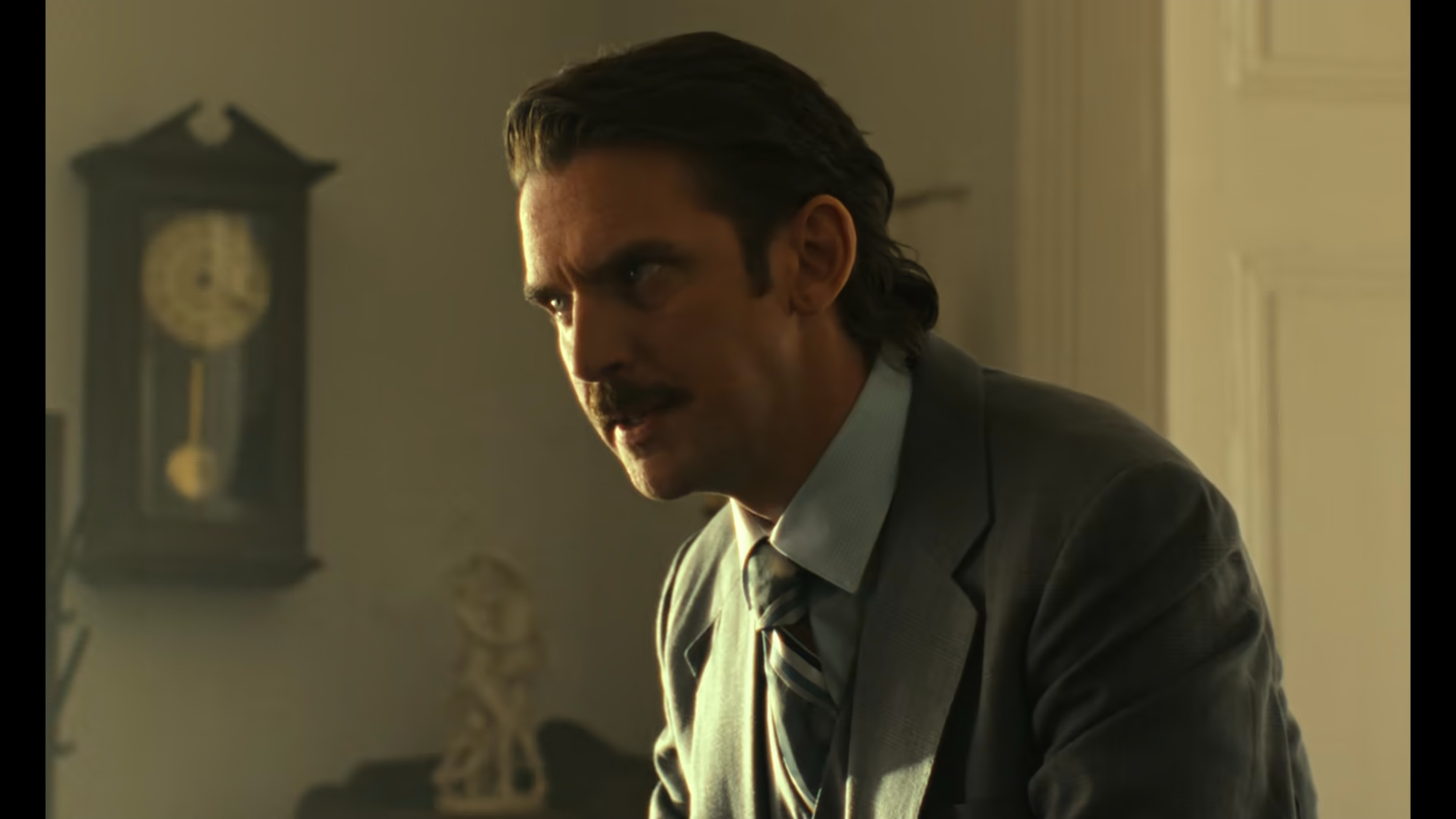 A man in a gray suit and tie is speaking indoors, with a vintage clock on the wall behind him.
