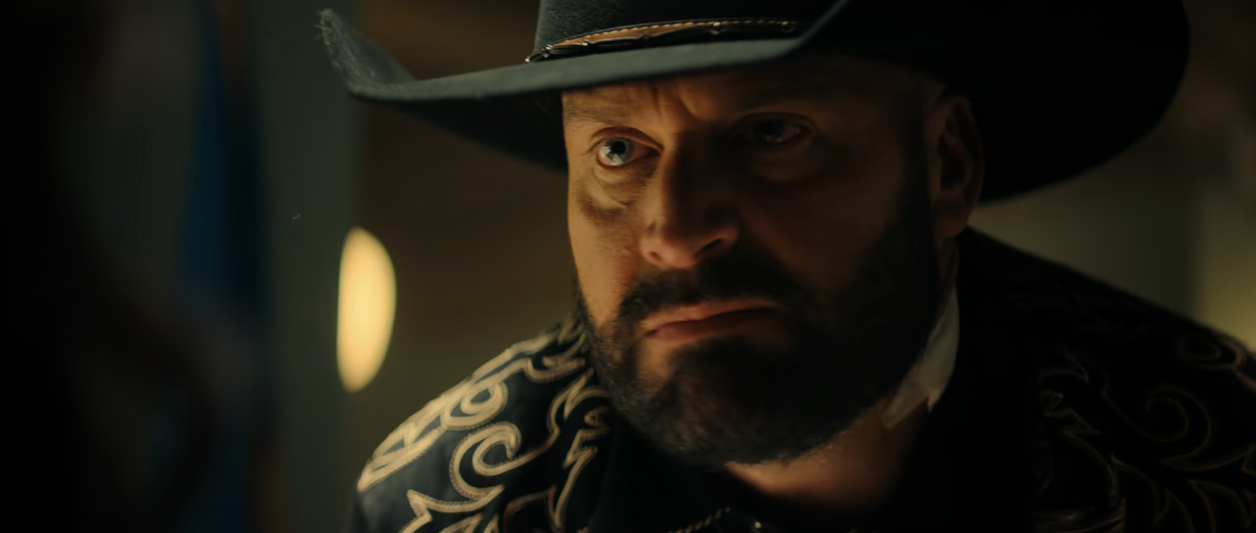 A man with a beard and mustache wearing a black cowboy hat with gold trim, a dark shirt with embroidered patterns, looking serious and intense in a dimly lit setting.