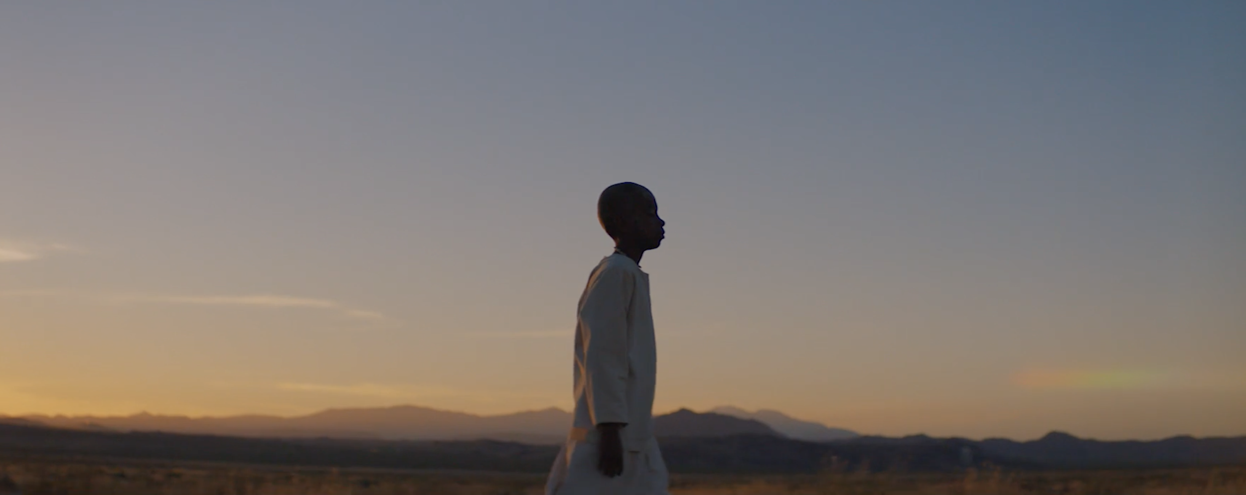 Silhouette of a person walking outdoors during sunset with mountains in the background.