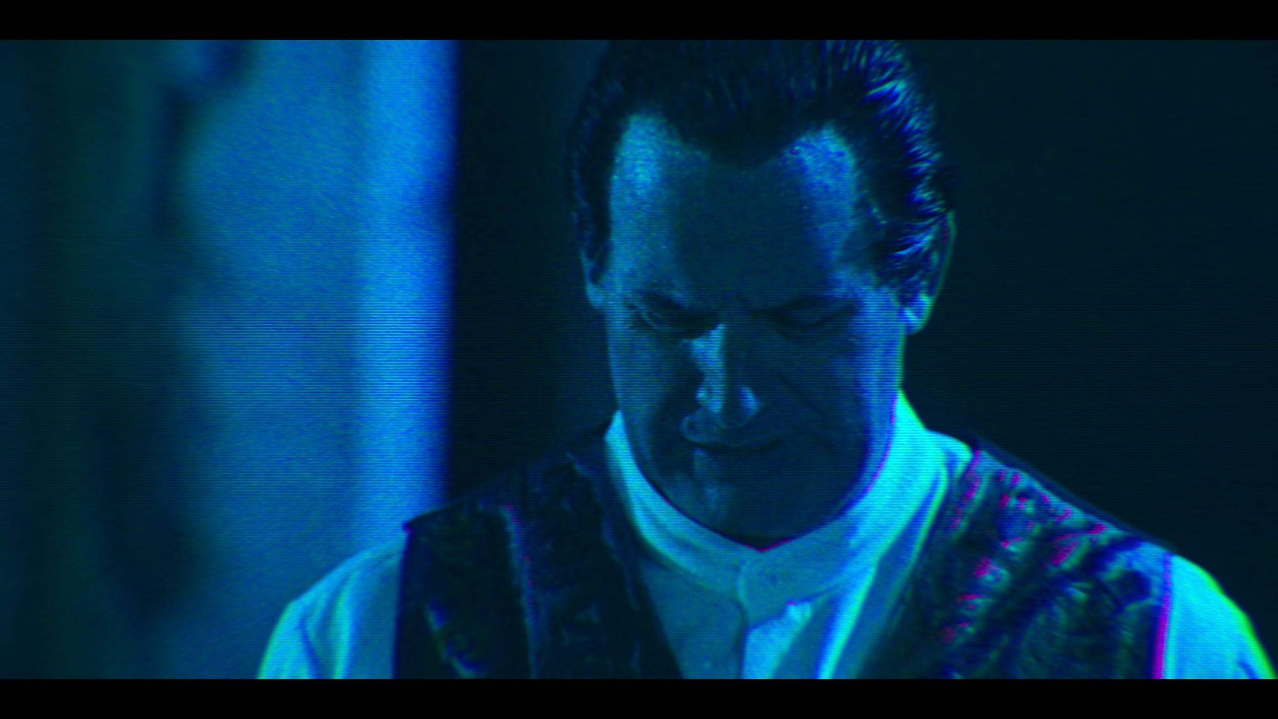 A man with slicked-back hair and a serious expression, dressed in a white shirt and dark vest, stands in a dark setting with a blue tint.