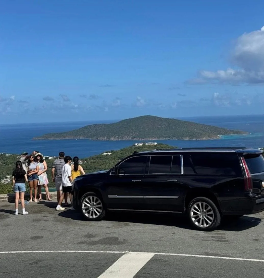 Tourists taking photos near a black limousine with an ocean and island view in the background on a clear day.