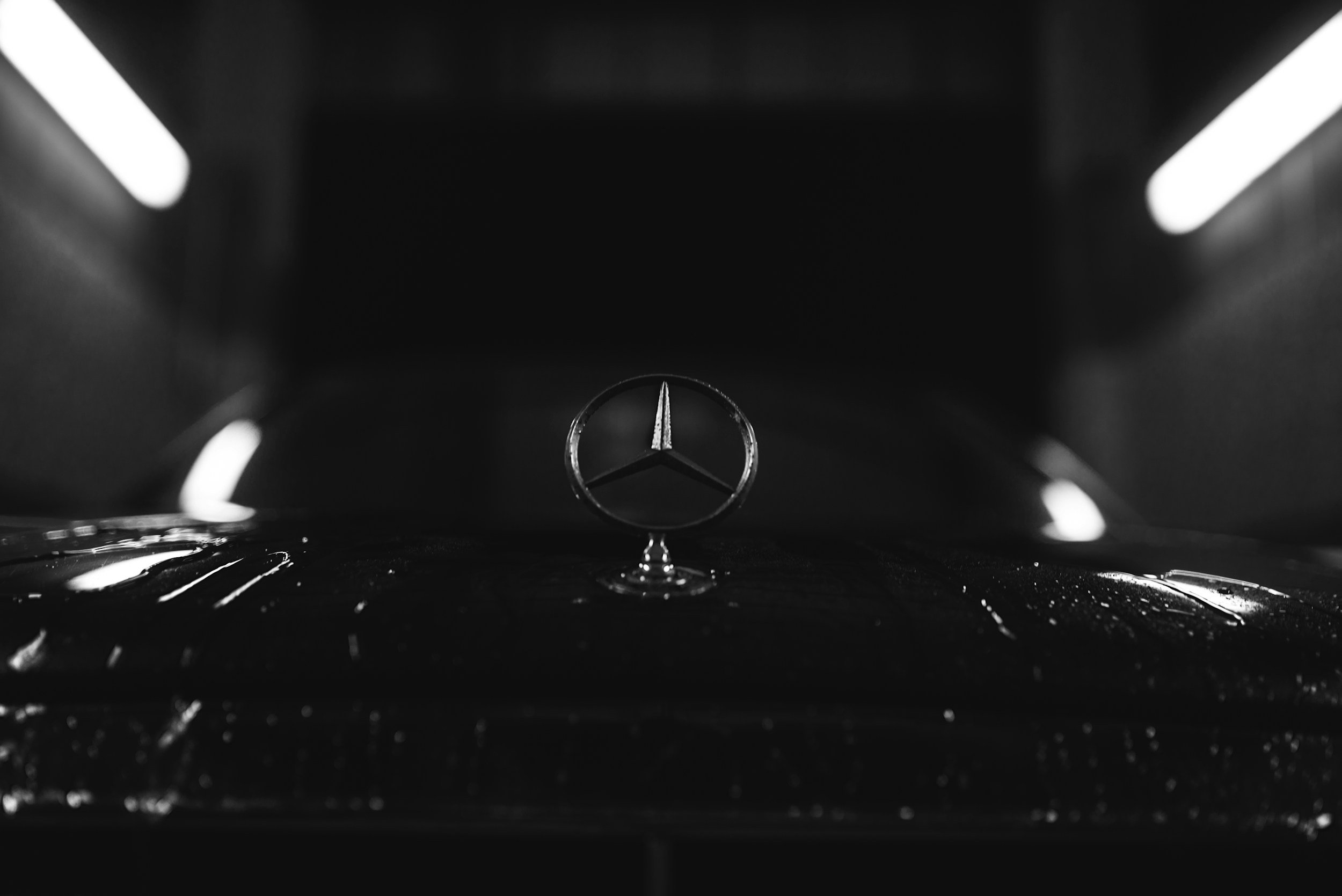 Close-up of a Mercedes-Benz emblem on the hood of a black car, with water droplets on the surface, in a dimly lit environment.