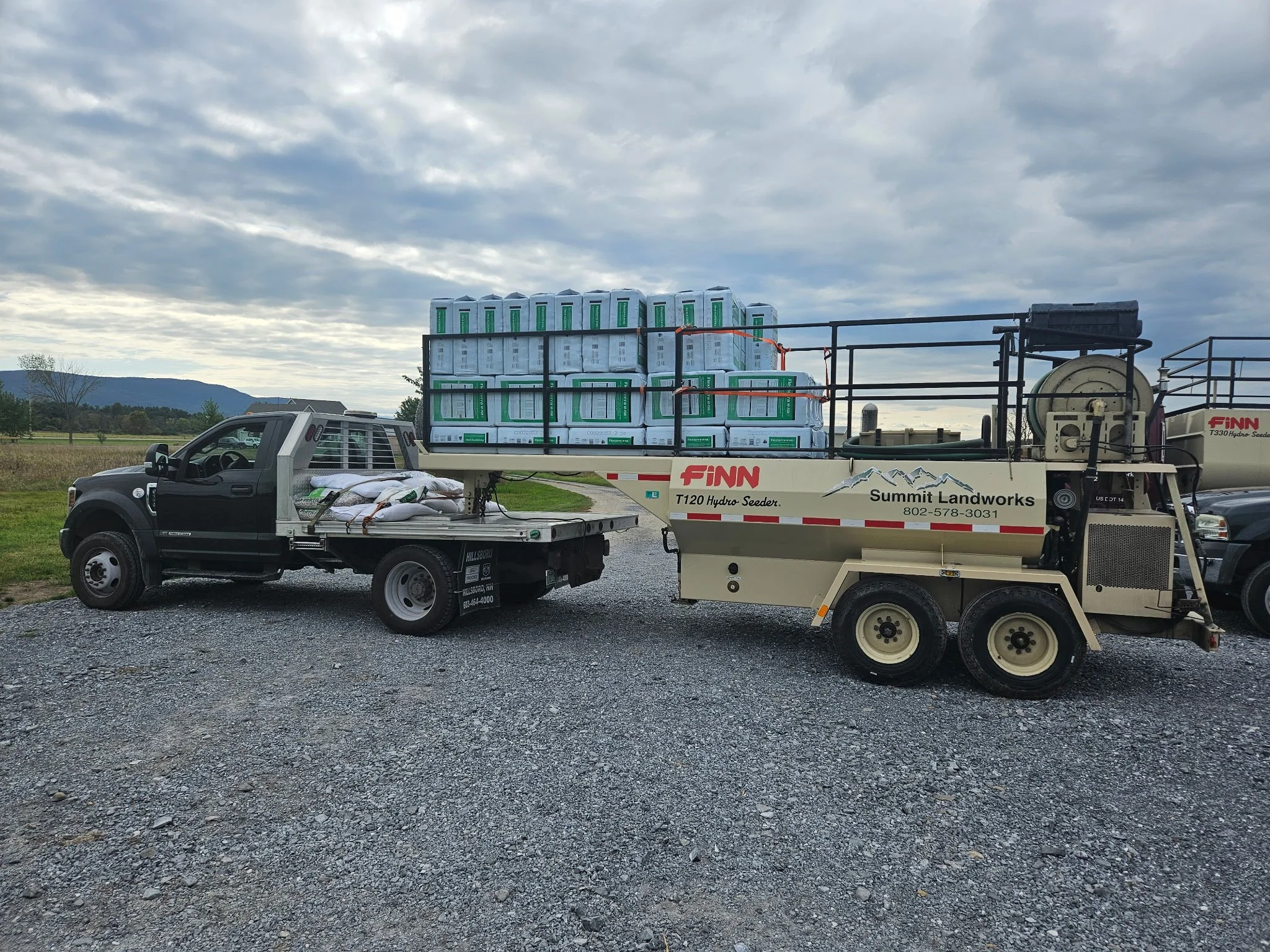 A utility truck and a hydro seeder trailer attached at an outdoor site, with stacks of bags and boxes on the trailer, and a cloudy sky overhead.