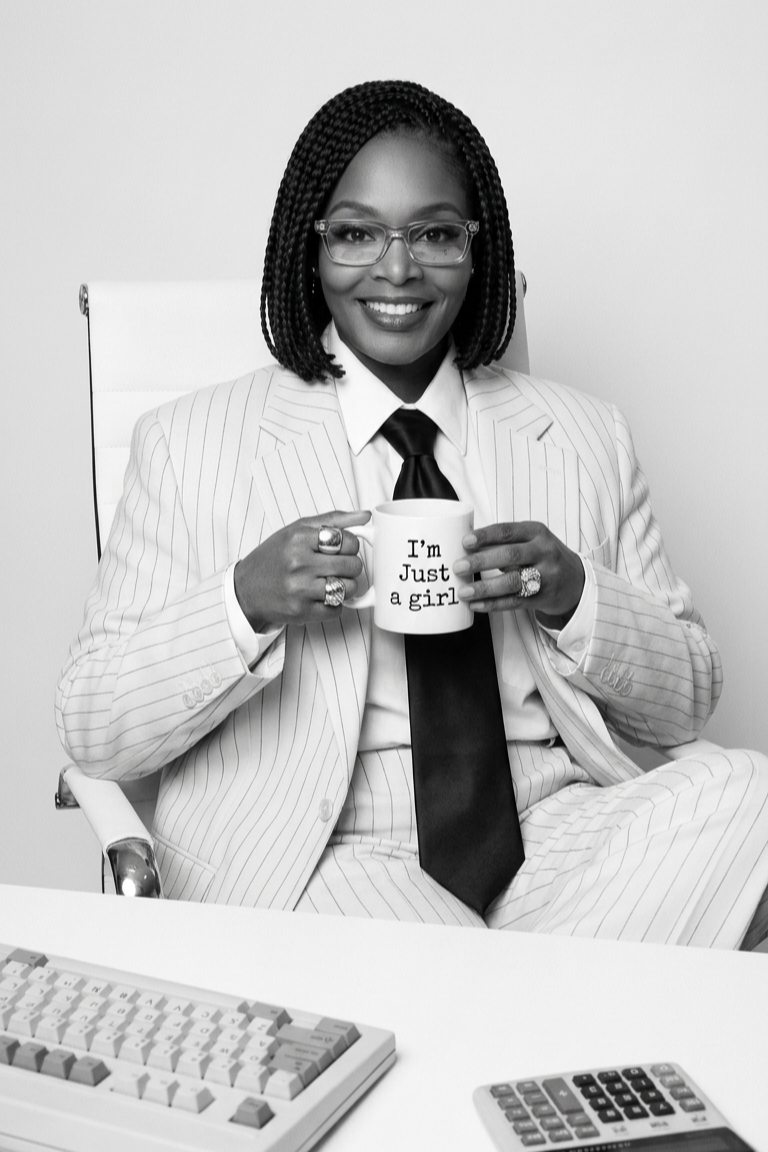 A woman in a pinstripe suit and glasses sitting on an office chair, smiling and holding a mug that says "I'm Just a girl" in an office setting.