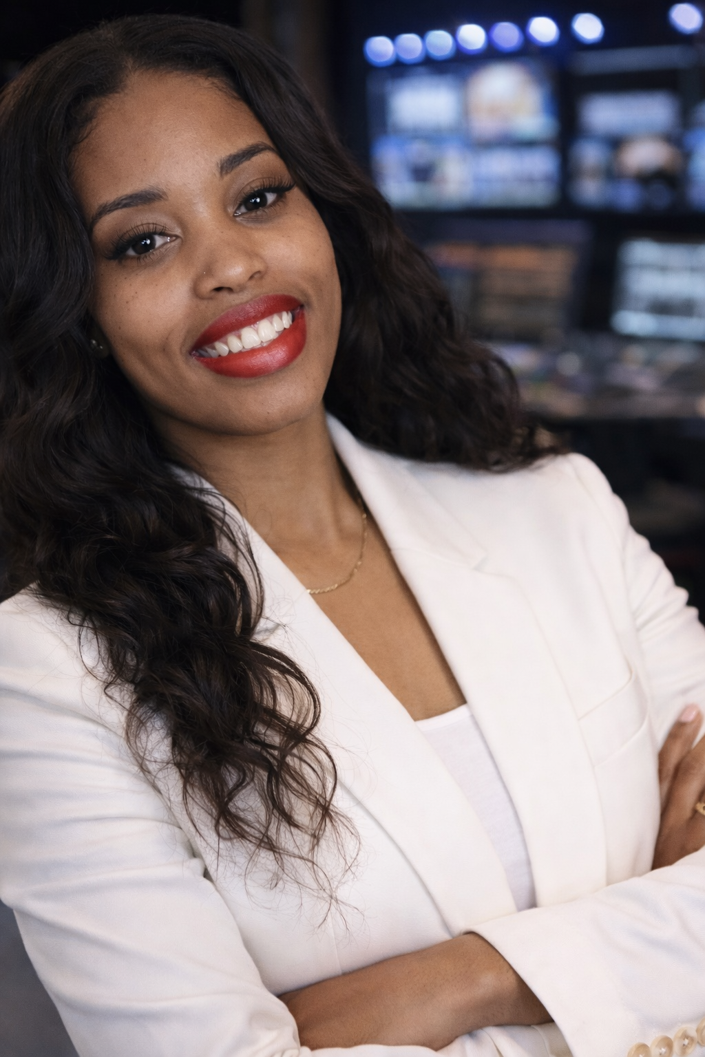 A woman with curly dark hair, red lipstick, white blazer, and crossed arms smiling at the camera in a professional setting.