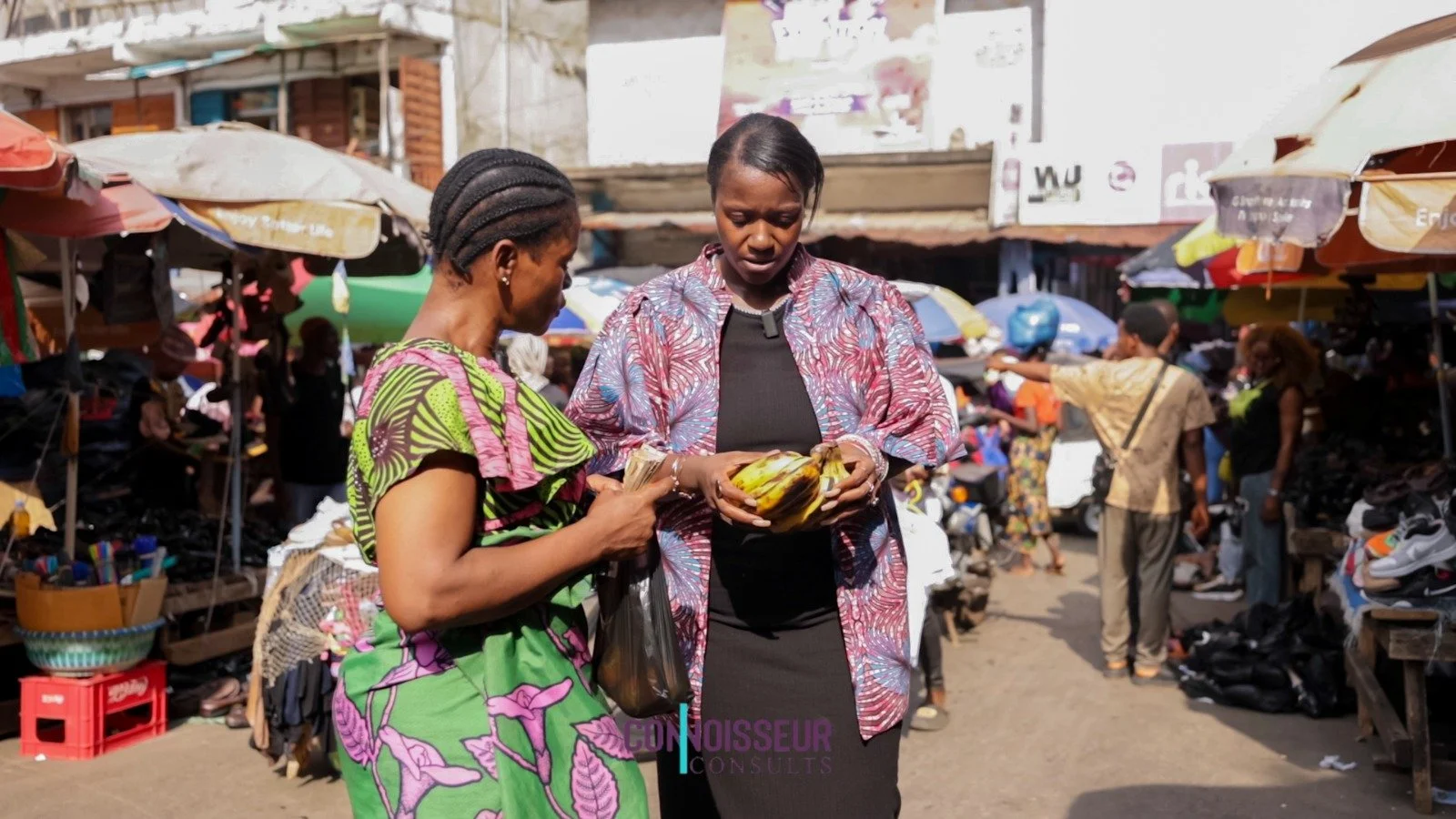 Two women, one in a colorful green and purple dress and the other in a patterned jacket and black dress, standing in a busy outdoor market, looking at a bundle of bananas.