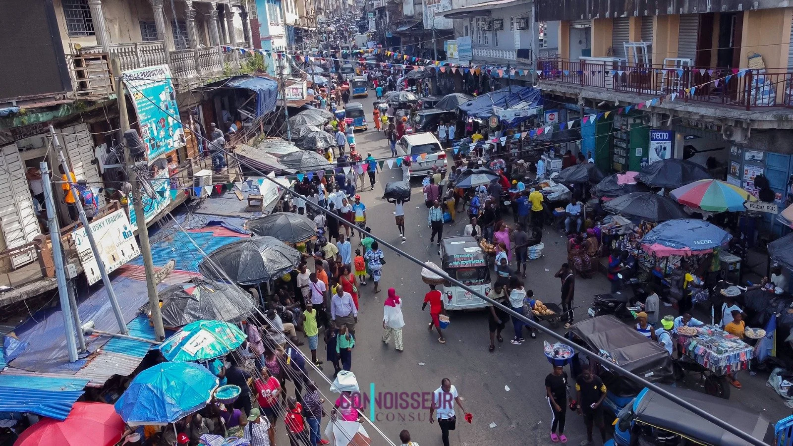 A busy street market with numerous vendors and shoppers. Stalls are covered with umbrellas and tarps. There are vehicles on the street and buildings on both sides.