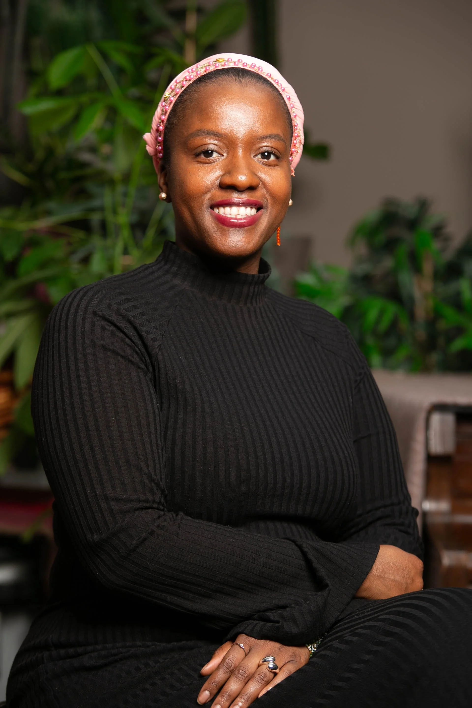 A woman with dark skin, wearing a black ribbed turtleneck, pink beaded headscarf, pearl earrings, and multiple rings, sitting indoors with green plants in the background, smiling at the camera.