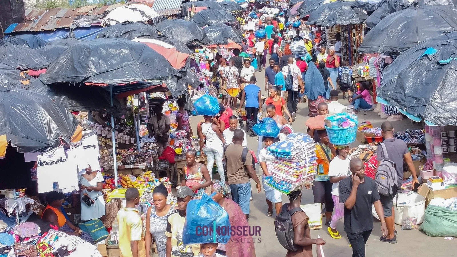 A busy outdoor market with vendors selling various goods under umbrellas, and many shoppers walking through the crowded street.