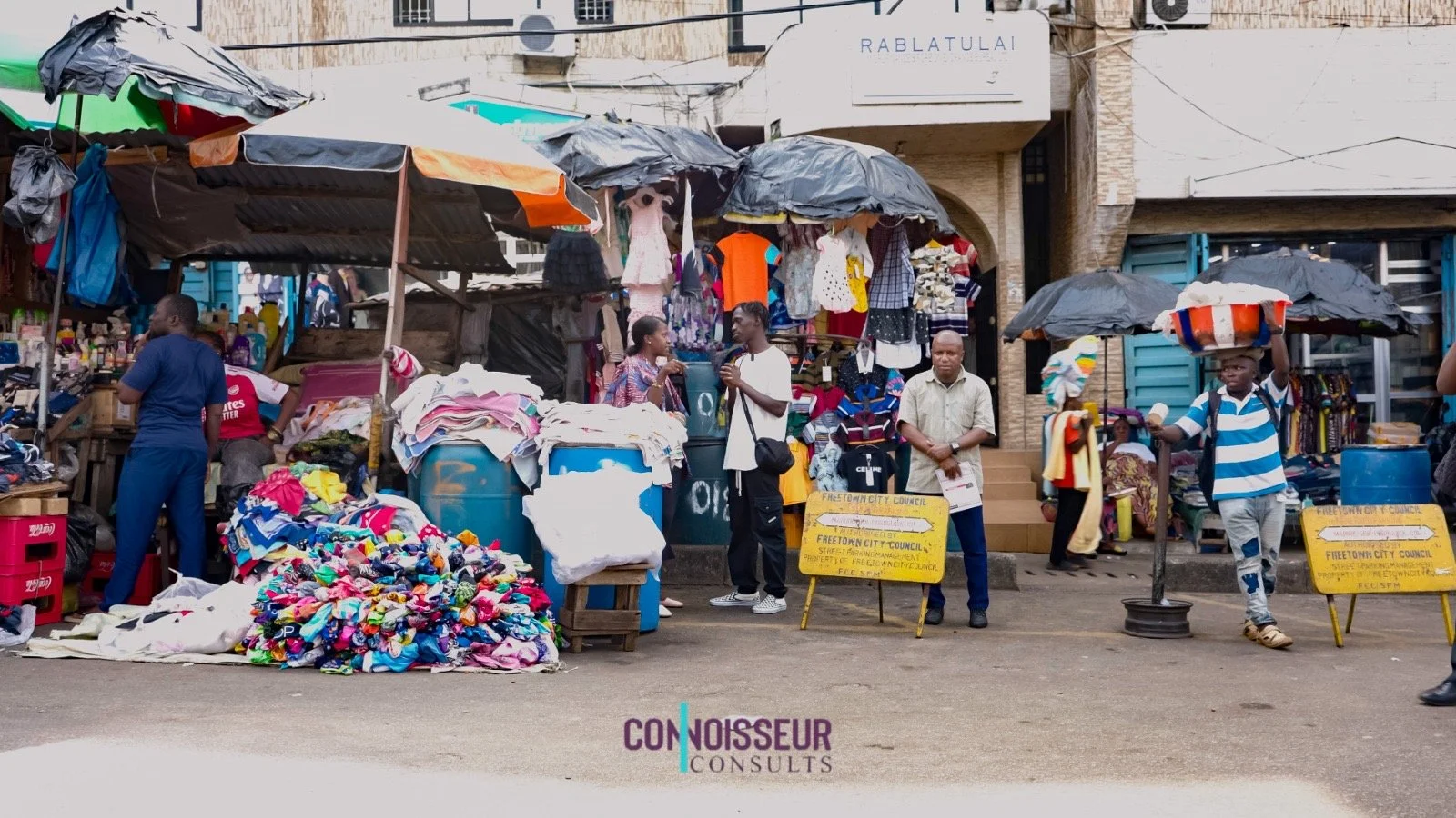 A busy outdoor market with vendors selling clothes and textiles under umbrellas, and several people walking or standing in front of the stalls on a street.
