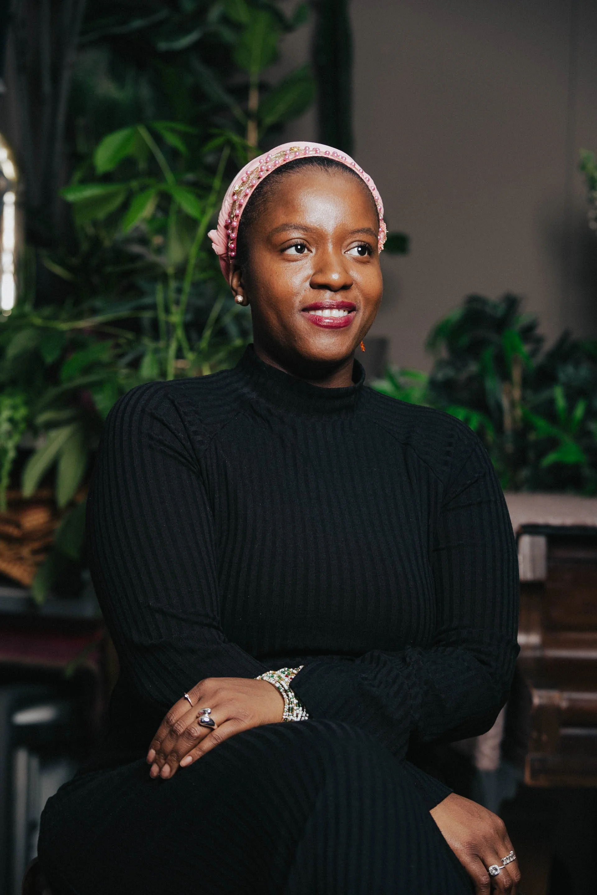 A woman with dark skin wearing a pink headscarf with pearls, a black ribbed top, and jewelry including rings, bracelets, and earrings. She is sitting with her arms crossed against a background of green plants and a dimly lit room.