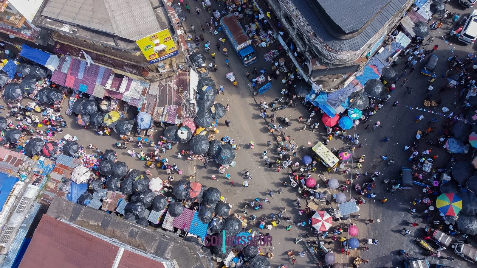 Aerial view of a busy street market with numerous vendors and shoppers, with umbrellas and makeshift stalls along the street.