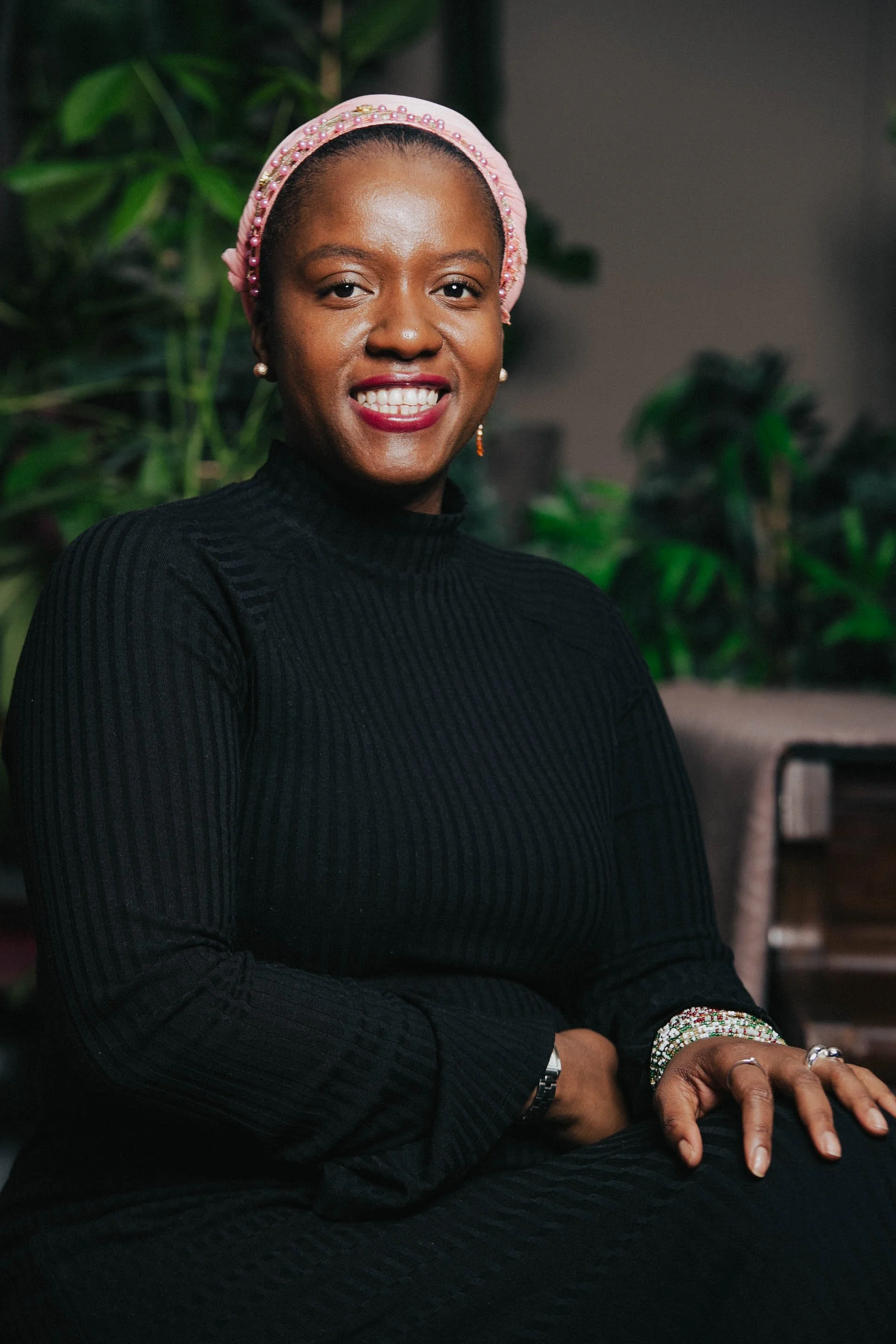 A woman with dark skin smiling, wearing a pink beaded headband, pearl earrings, a black ribbed turtleneck, and a beaded bracelet, seated indoors with green plants in the background.