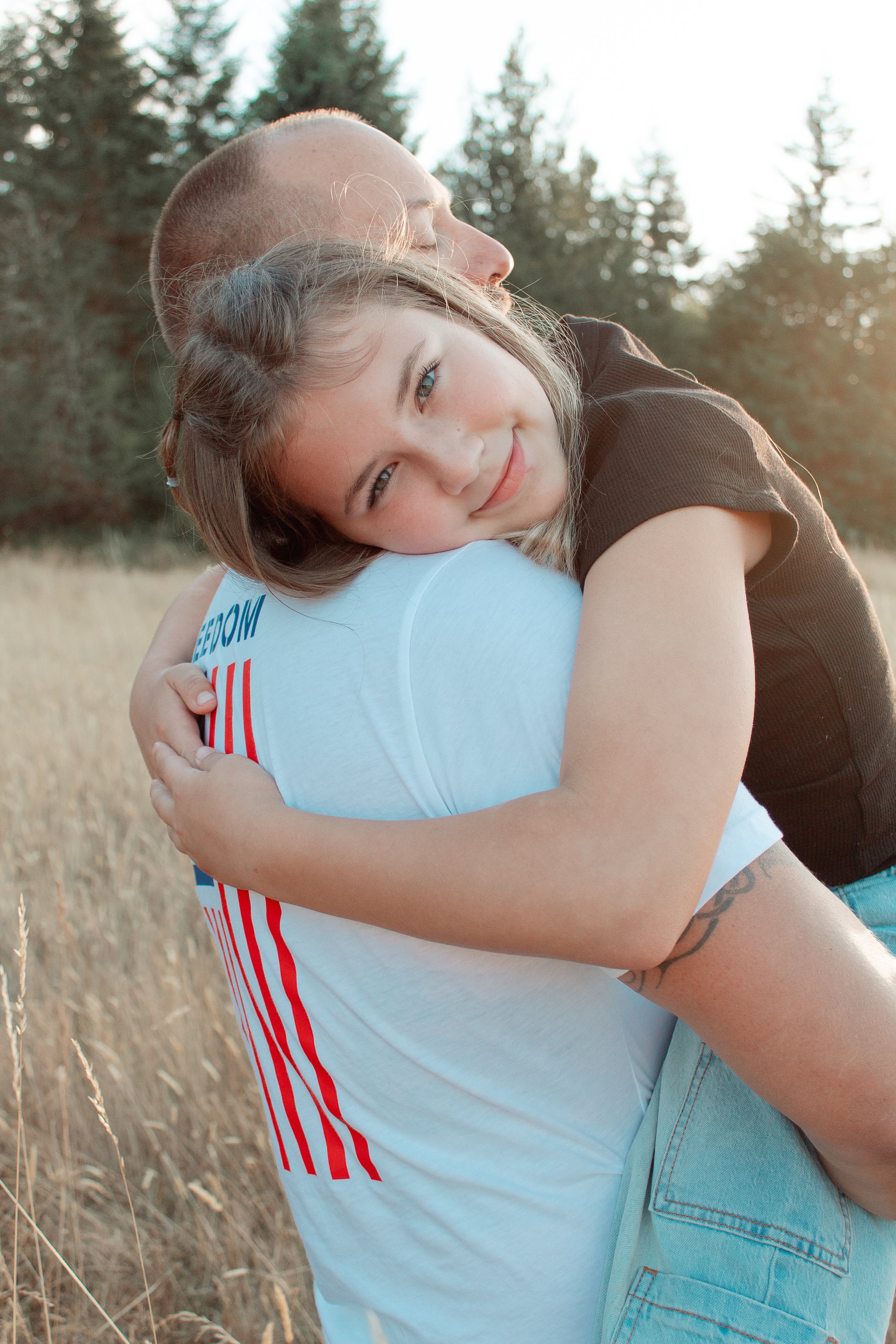 A young girl with brown hair and a slight smile is being embraced by a man in an outdoor setting with trees and tall grass, during what appears to be late afternoon or early evening.