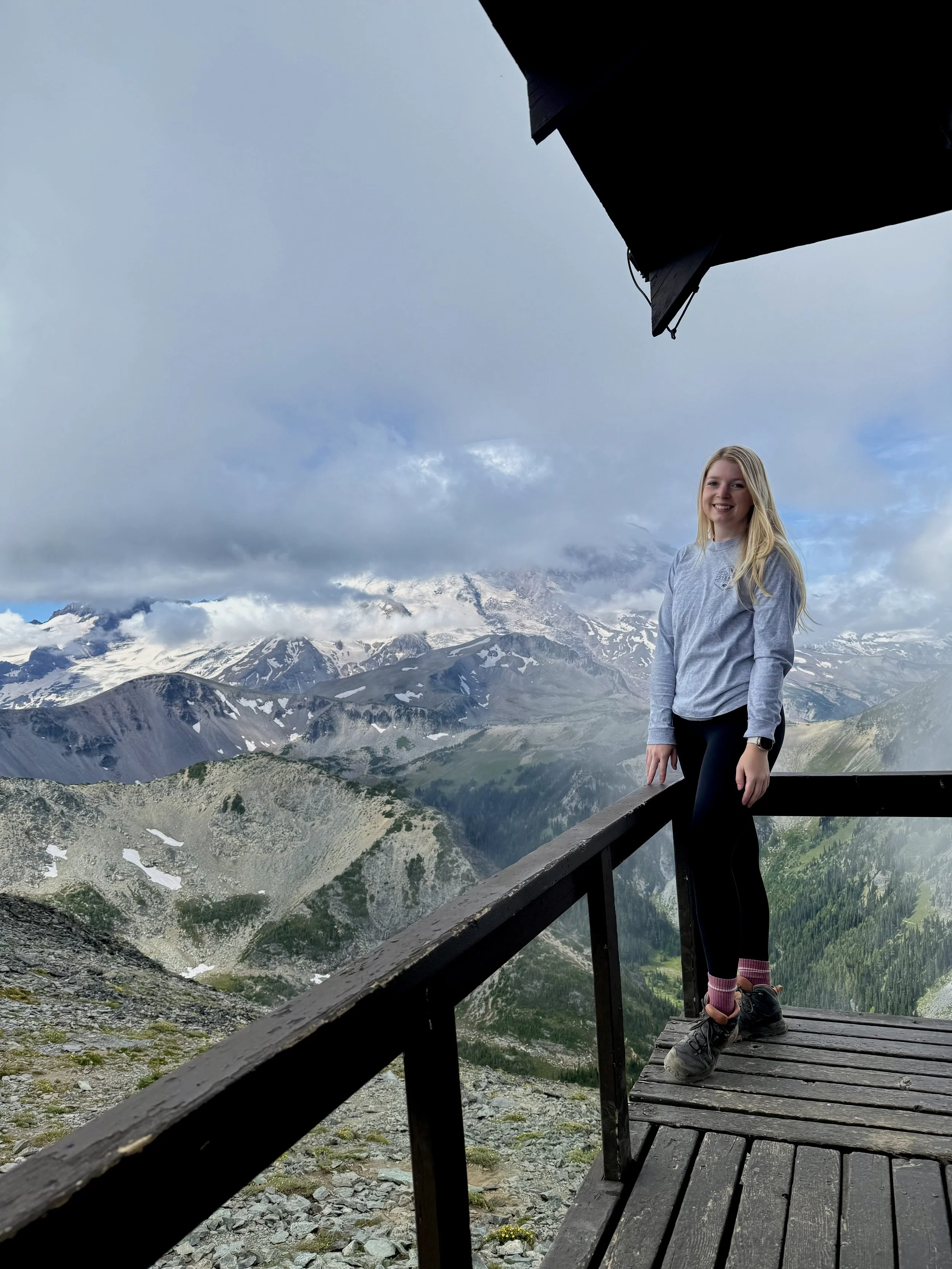 A woman standing on a wooden observation deck on a mountain, with snow-capped peaks and a cloudy sky in the background.