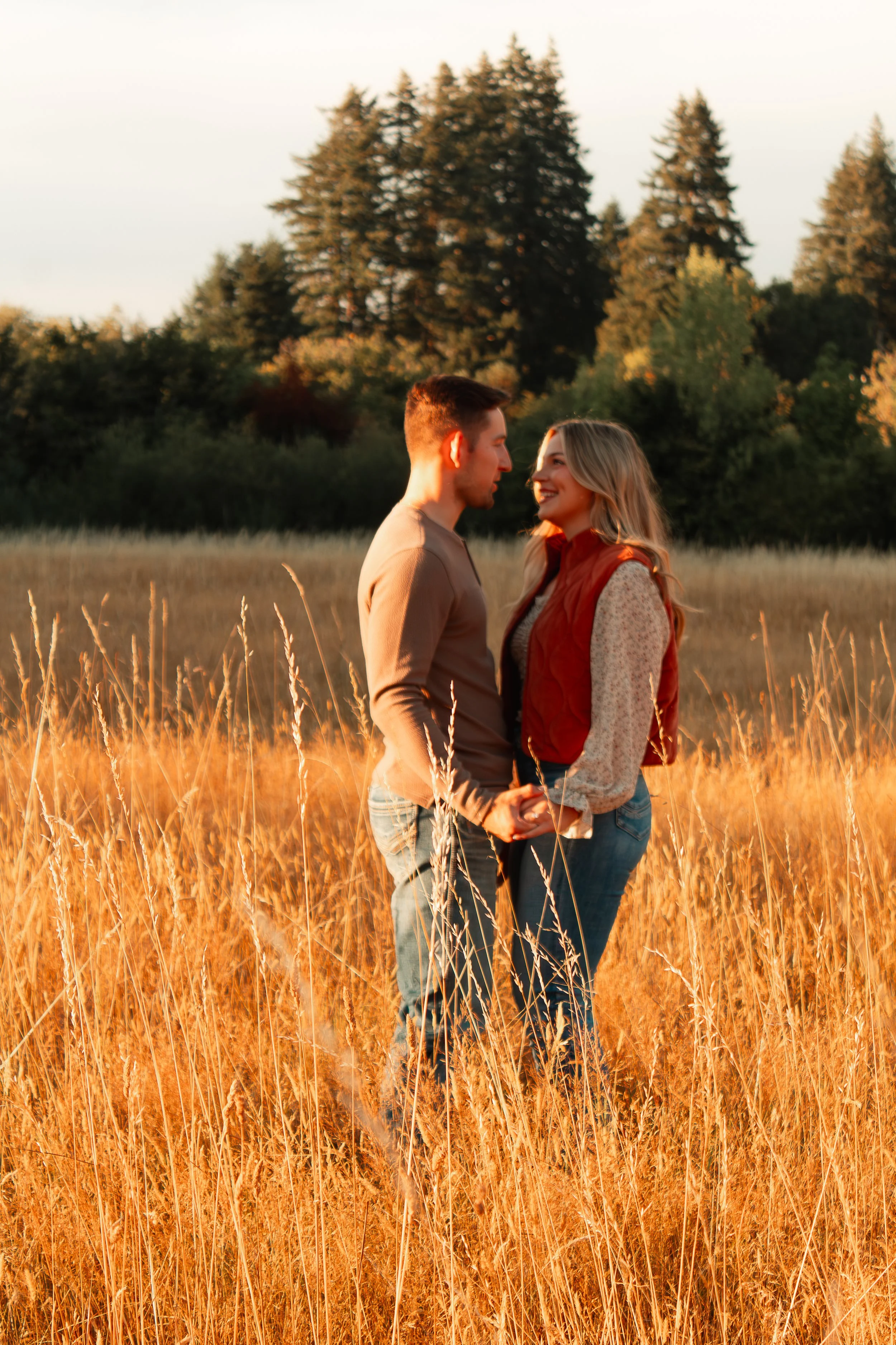A couple holding hands and smiling at each other in a golden field during sunset, with trees in the background.