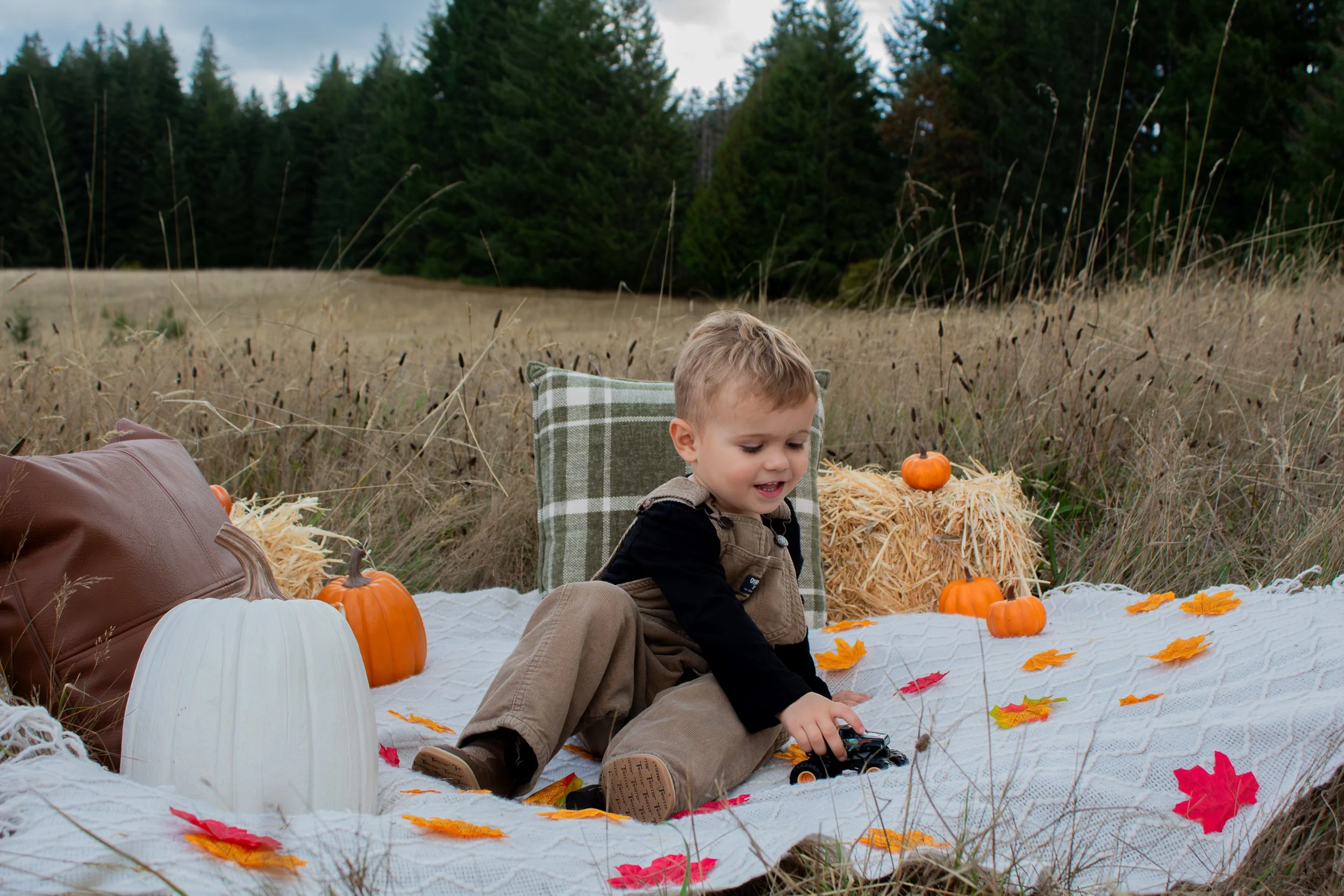 A young boy sitting on a white blanket outdoors in a field, surrounded by pumpkins, hay bales, and fall leaves.