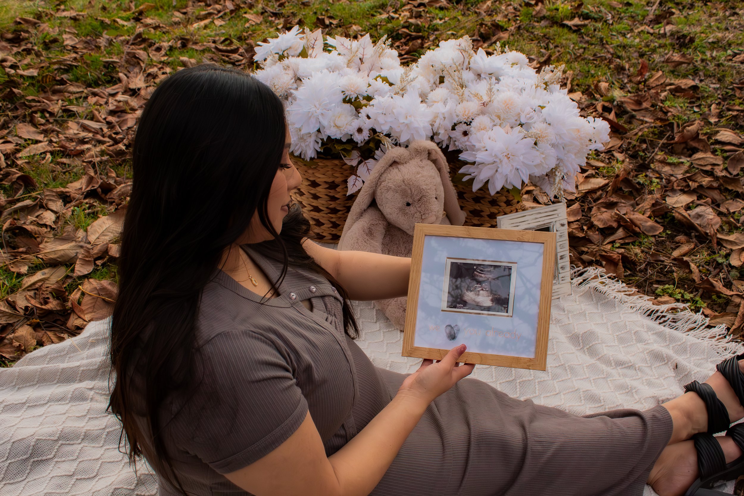 A woman is sitting on a blanket outdoors, holding a framed ultrasound picture, with a basket of white flowers and a plush bunny toy nearby, for a pregnancy announcement or memorial.