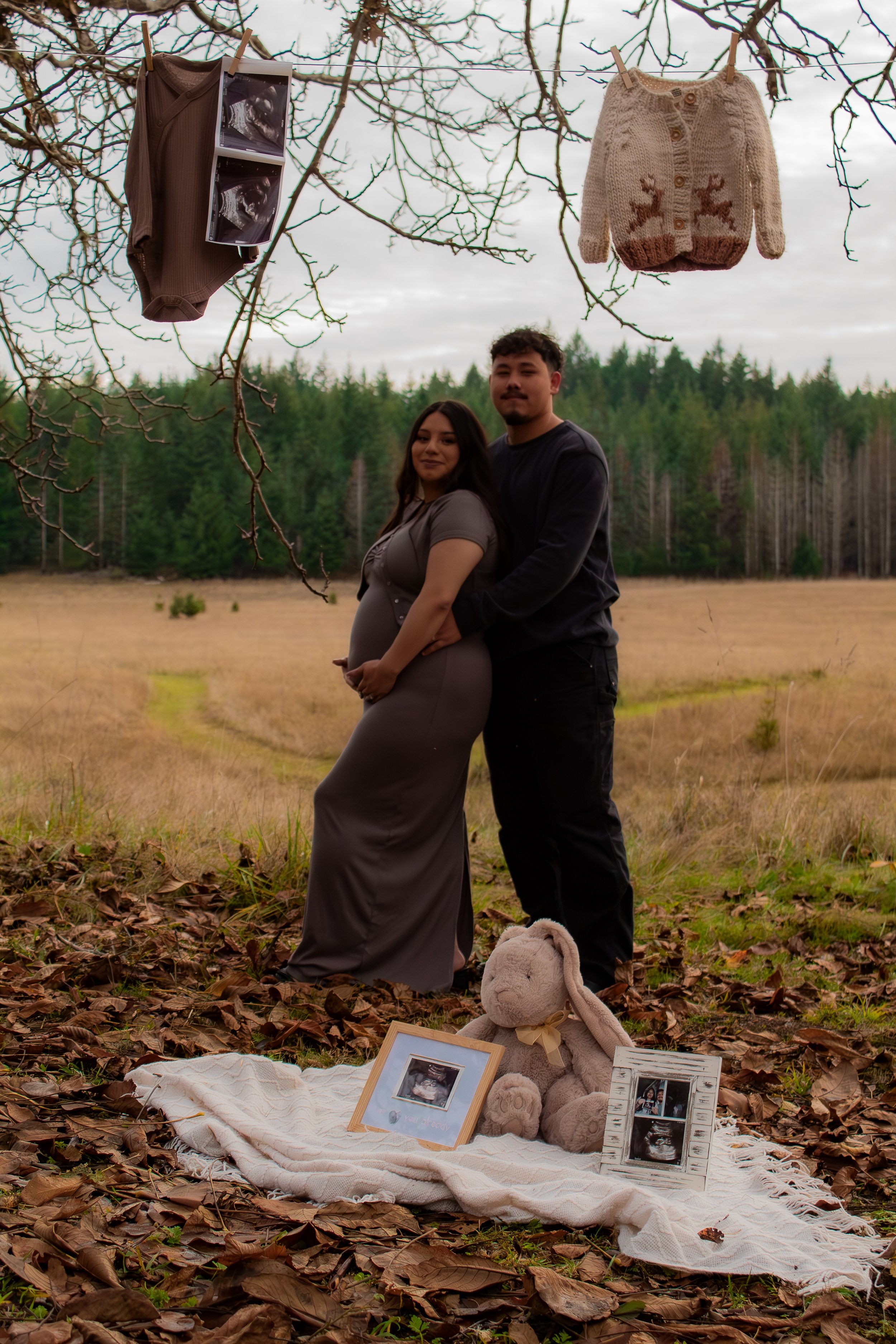 A couple, with a pregnant woman, standing outdoors in a field with trees in the background, holding hands and smiling at the camera. Clothes, ultrasound photos, a framed picture, a stuffed bunny, and baby items hang from a tree overhead, and a blanke