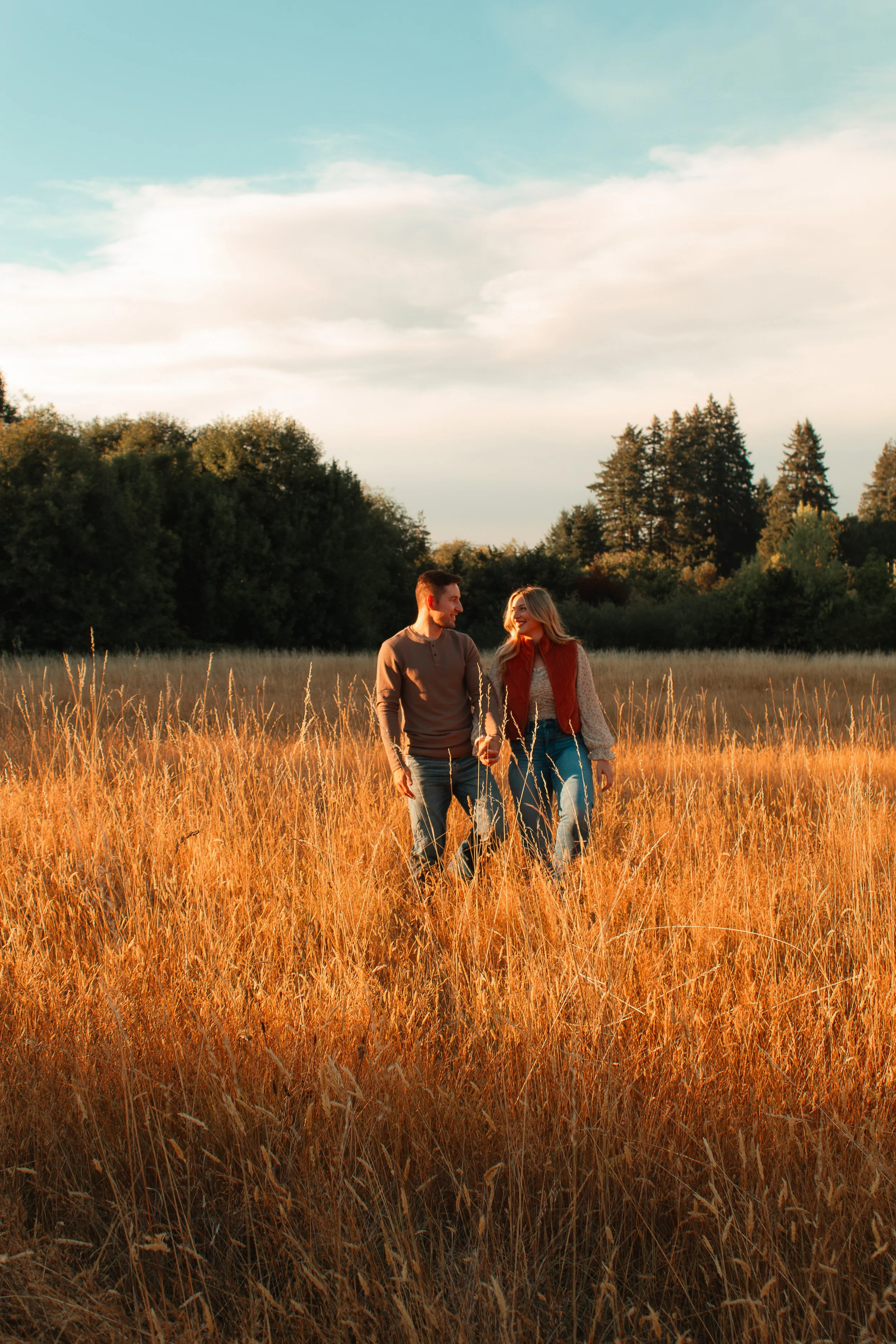 A couple walking hand in hand through a golden field at sunset, surrounded by trees with a partly cloudy sky above.