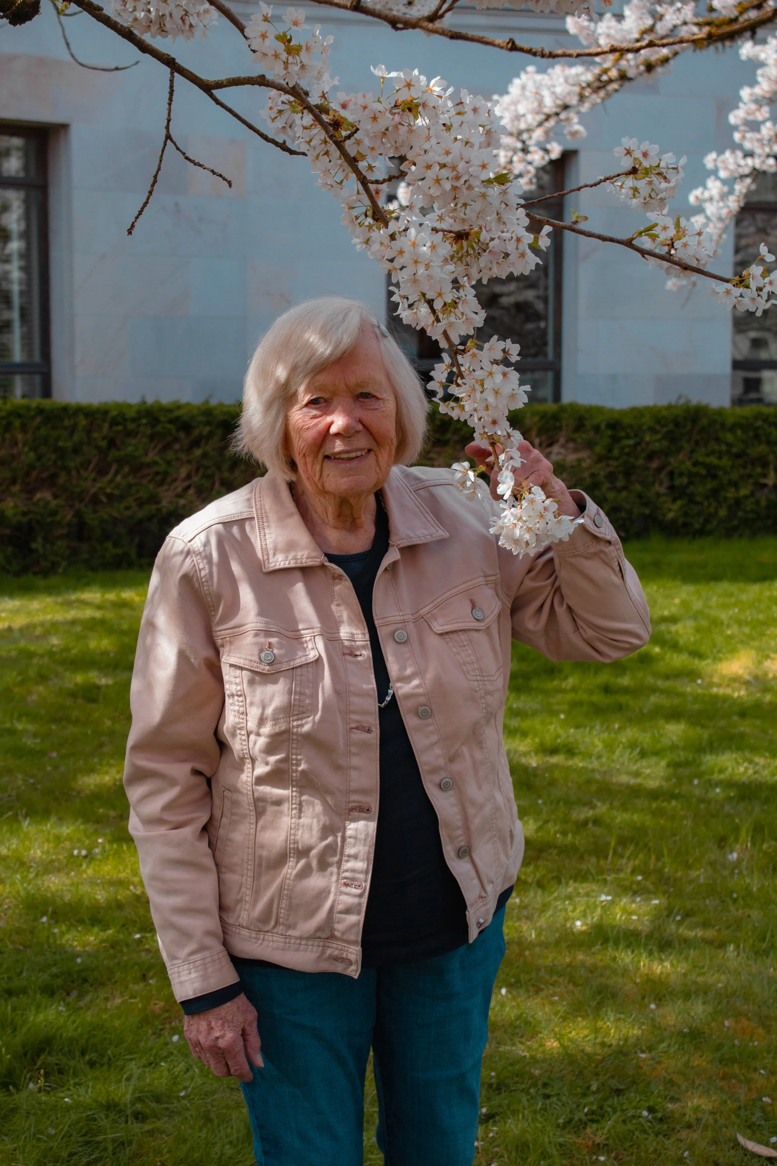 An elderly woman with blonde hair, wearing a light pink jacket and blue jeans, standing outdoors under a blooming cherry blossom tree. She is smiling and holding a branch of cherry blossoms.