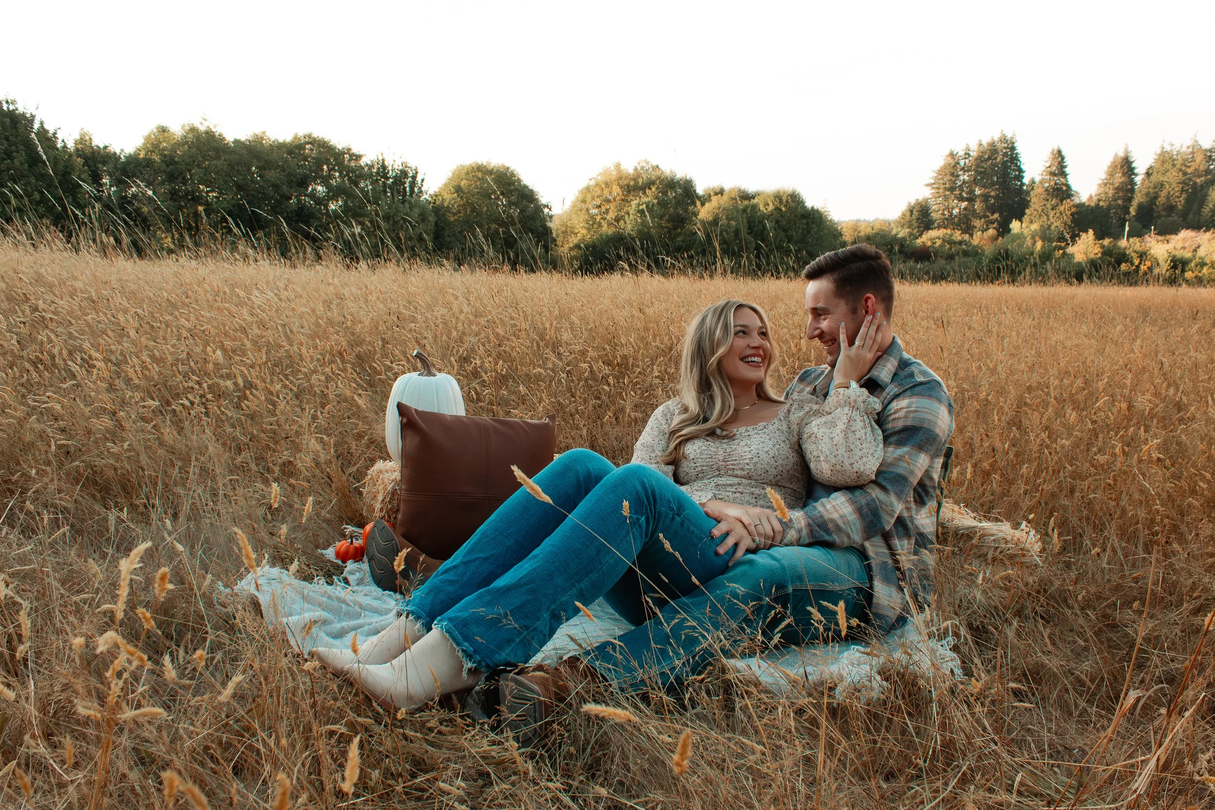 A happy couple sitting on a blanket in a golden field, smiling at each other during fall, with a pumpkin and pillow nearby.