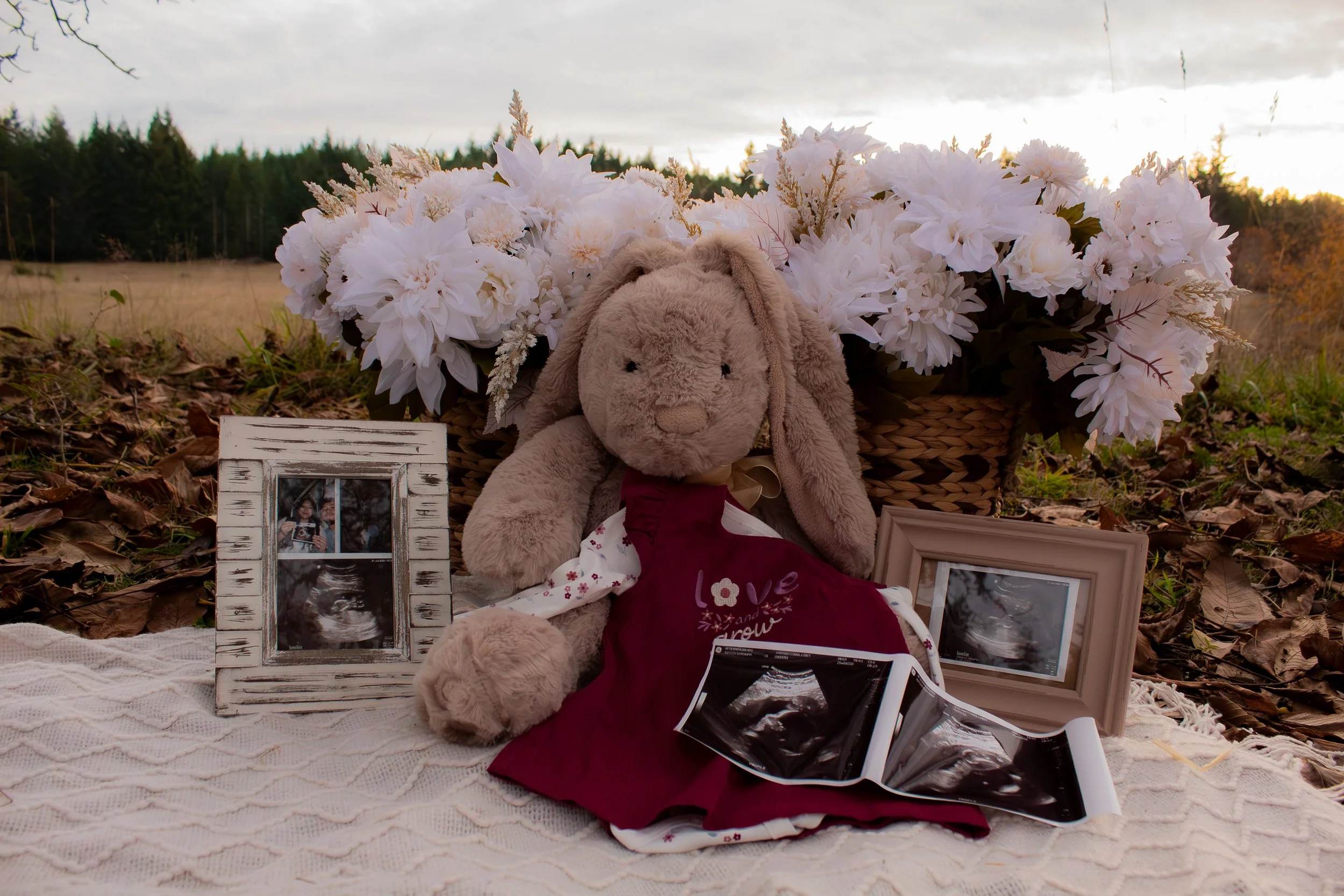 A memorial display outdoors with a plush bunny toy, framed ultrasound photos, a photo of a baby, a basket of white flowers, and stray leaves on the ground.