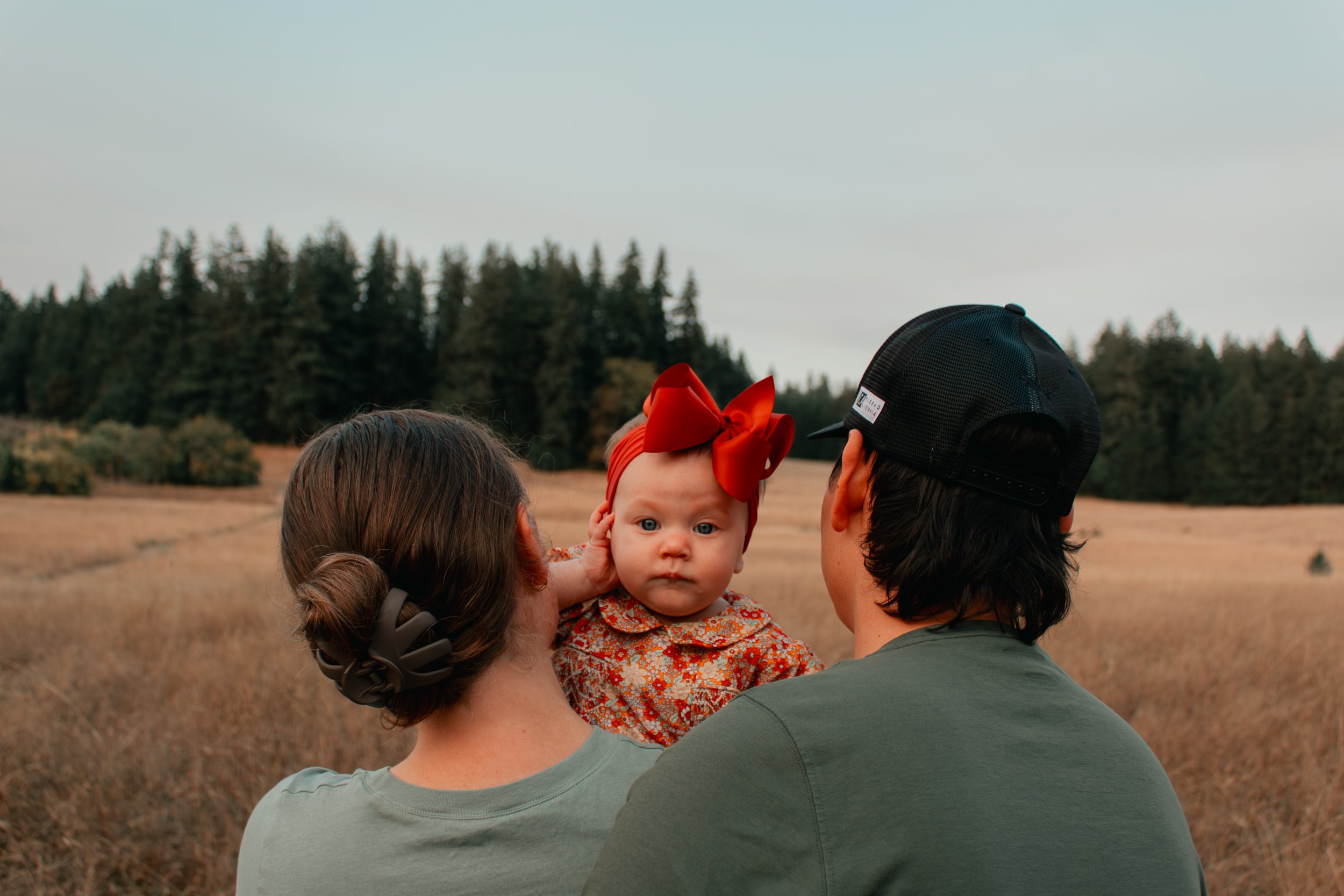 A family of three, a woman, a man, and a baby girl with a red bow, outdoors in a field with trees in the background.