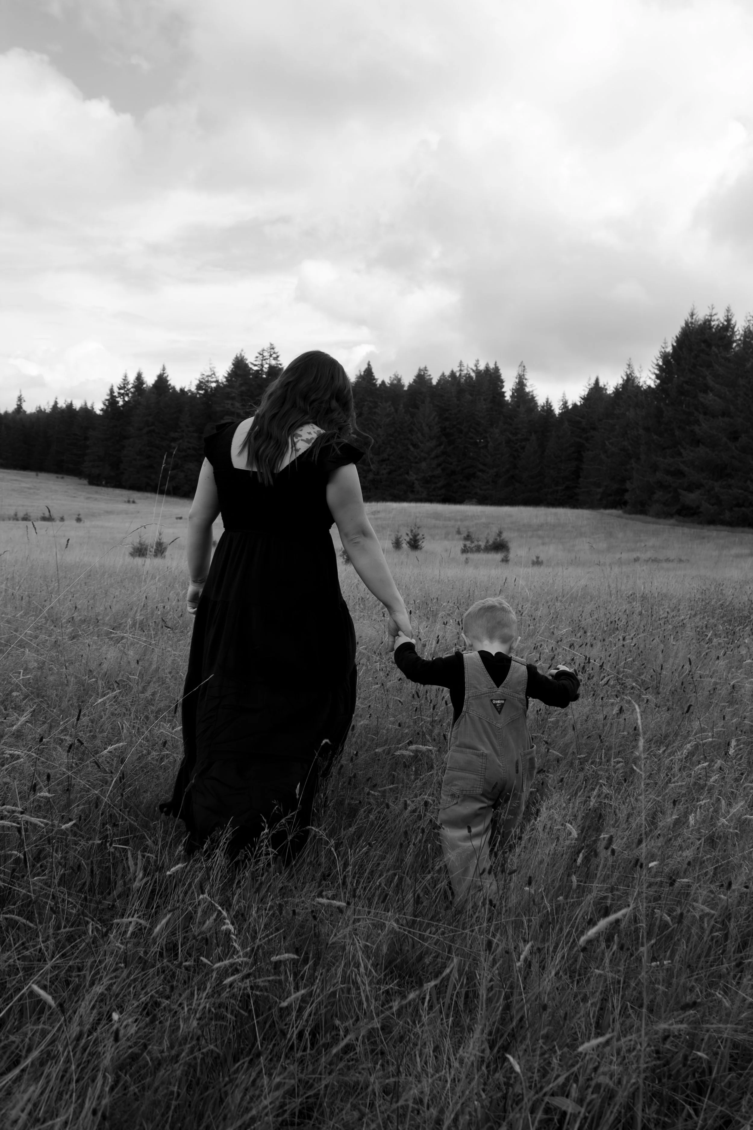 A woman and a young child walking hand in hand through a grassy field, with trees and clouds in the background, in black and white.